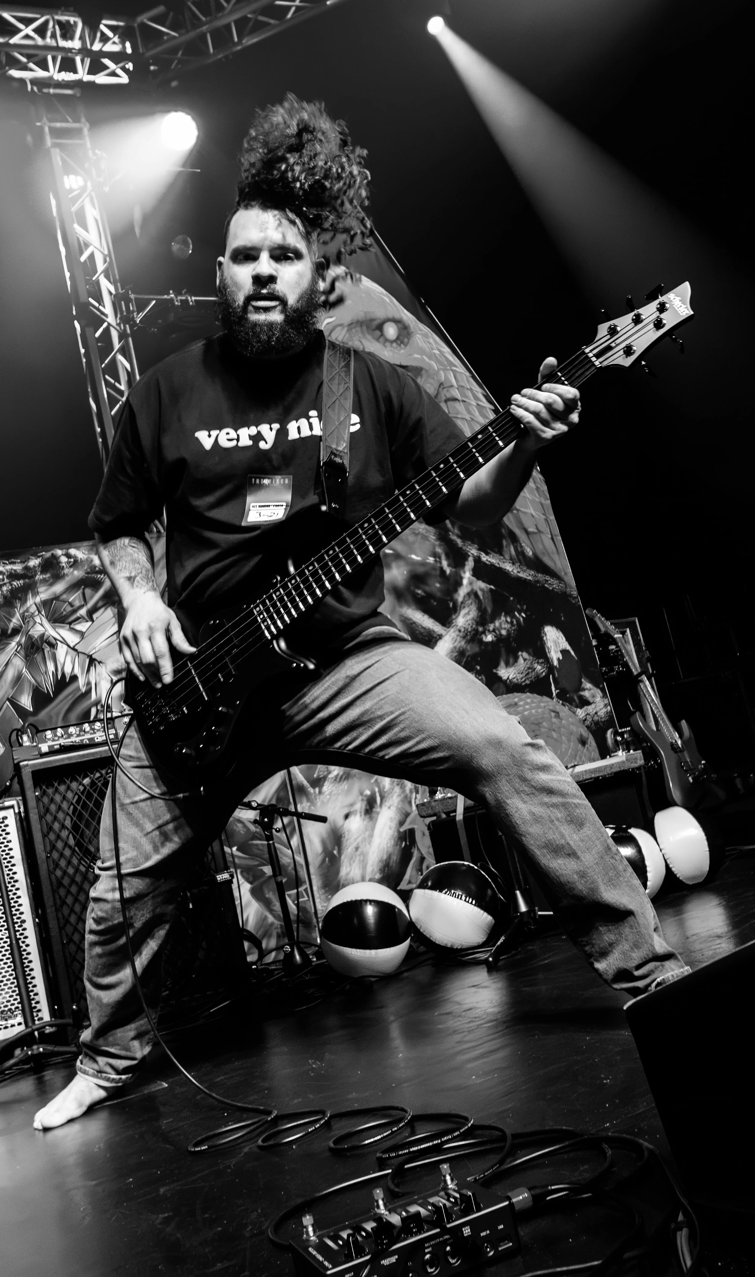 A man with curly hair in a hairdo and a beard playing an electric bass guitar on stage, with stage lights and equipment around him. He is wearing a T-shirt that says 'very nice' and appears to be performing energetically.