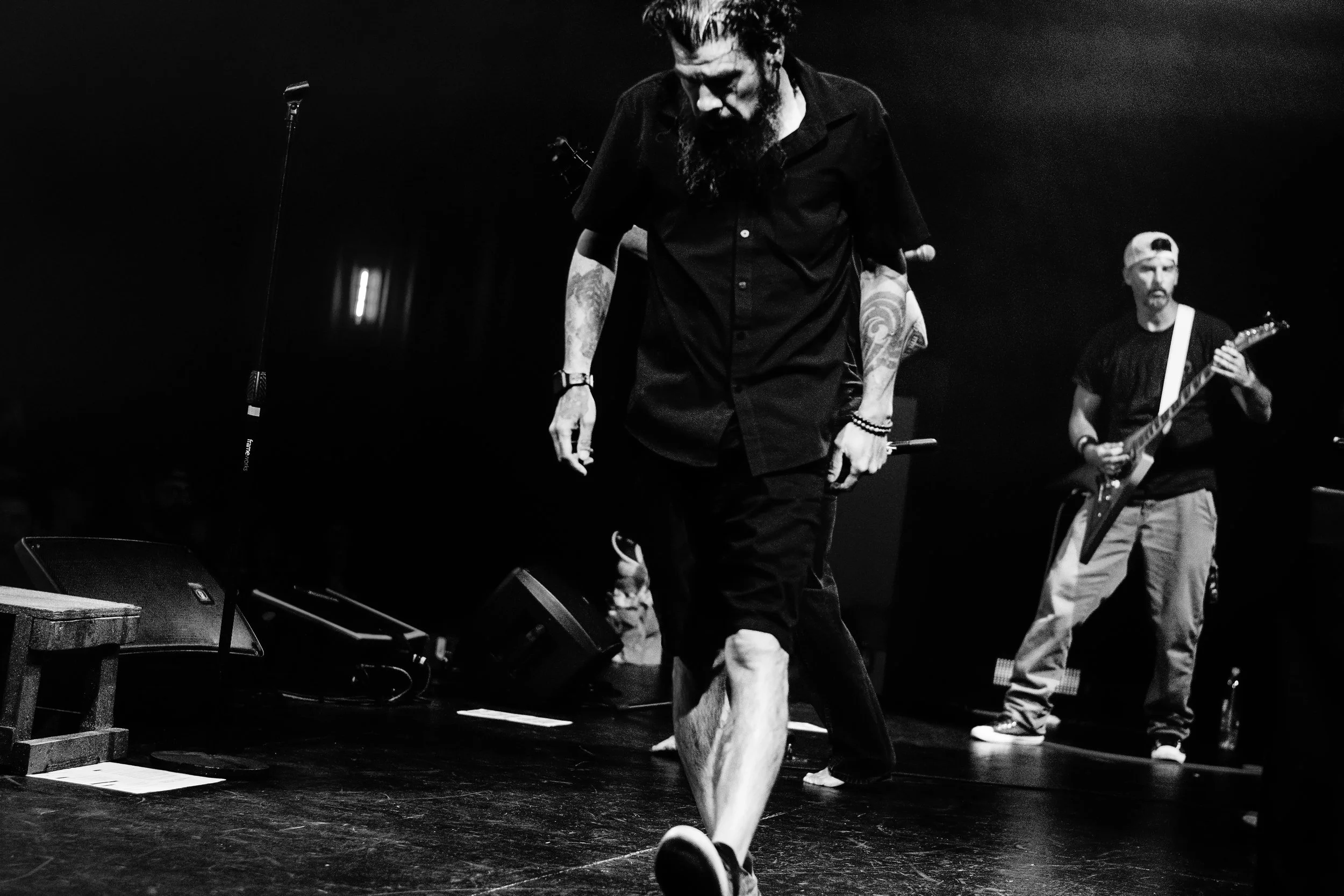 A black and white photo of a man with tattoos on his arms, a beard, and with hair tied back, walking barefoot on stage. In the background, another person is playing an electric guitar. Stage monitors and a microphone stand are visible.