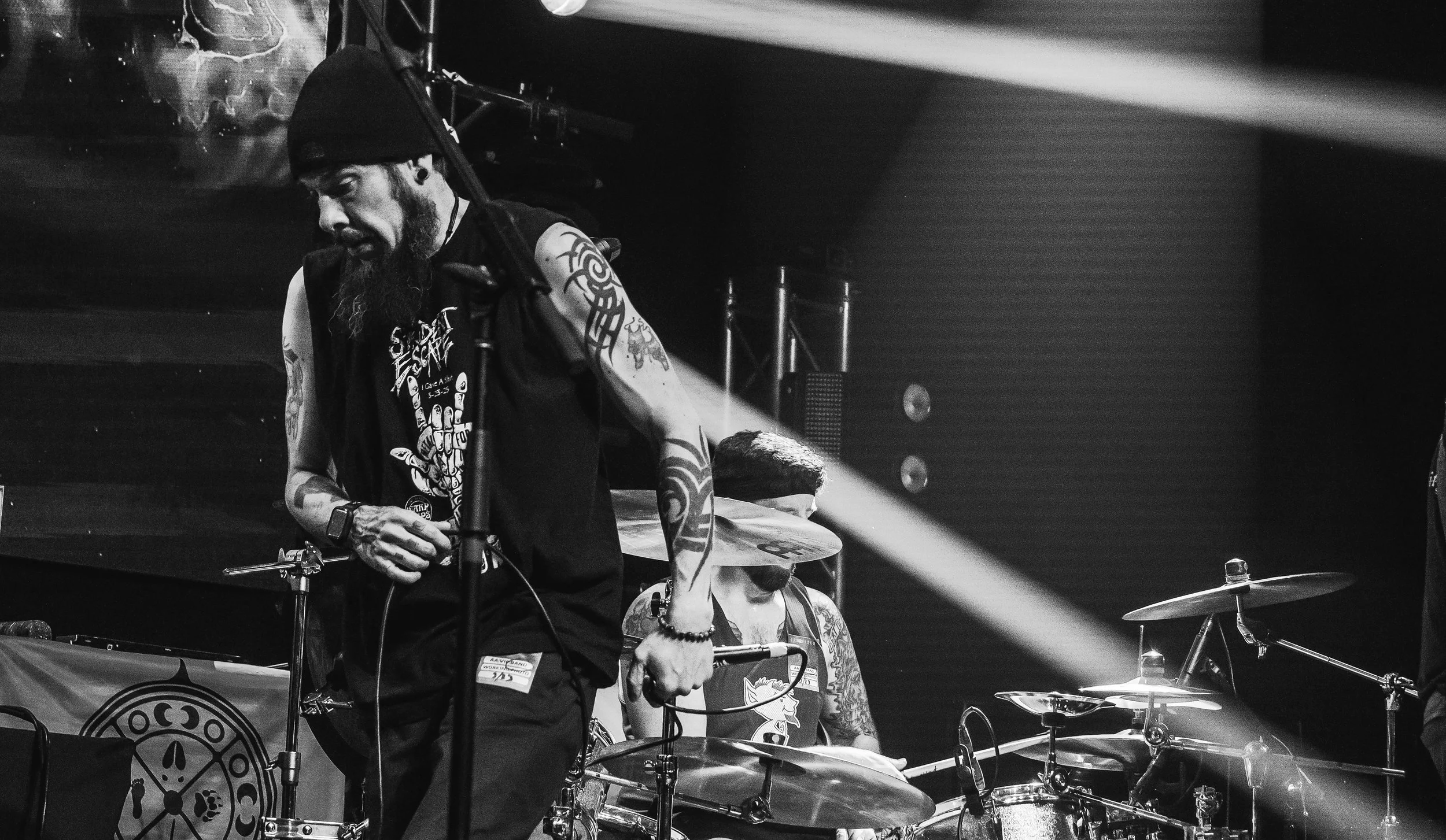 Black and white photo of a band performing on stage, showing a drummer and a musician with tattoos, a beard, and a beanie.