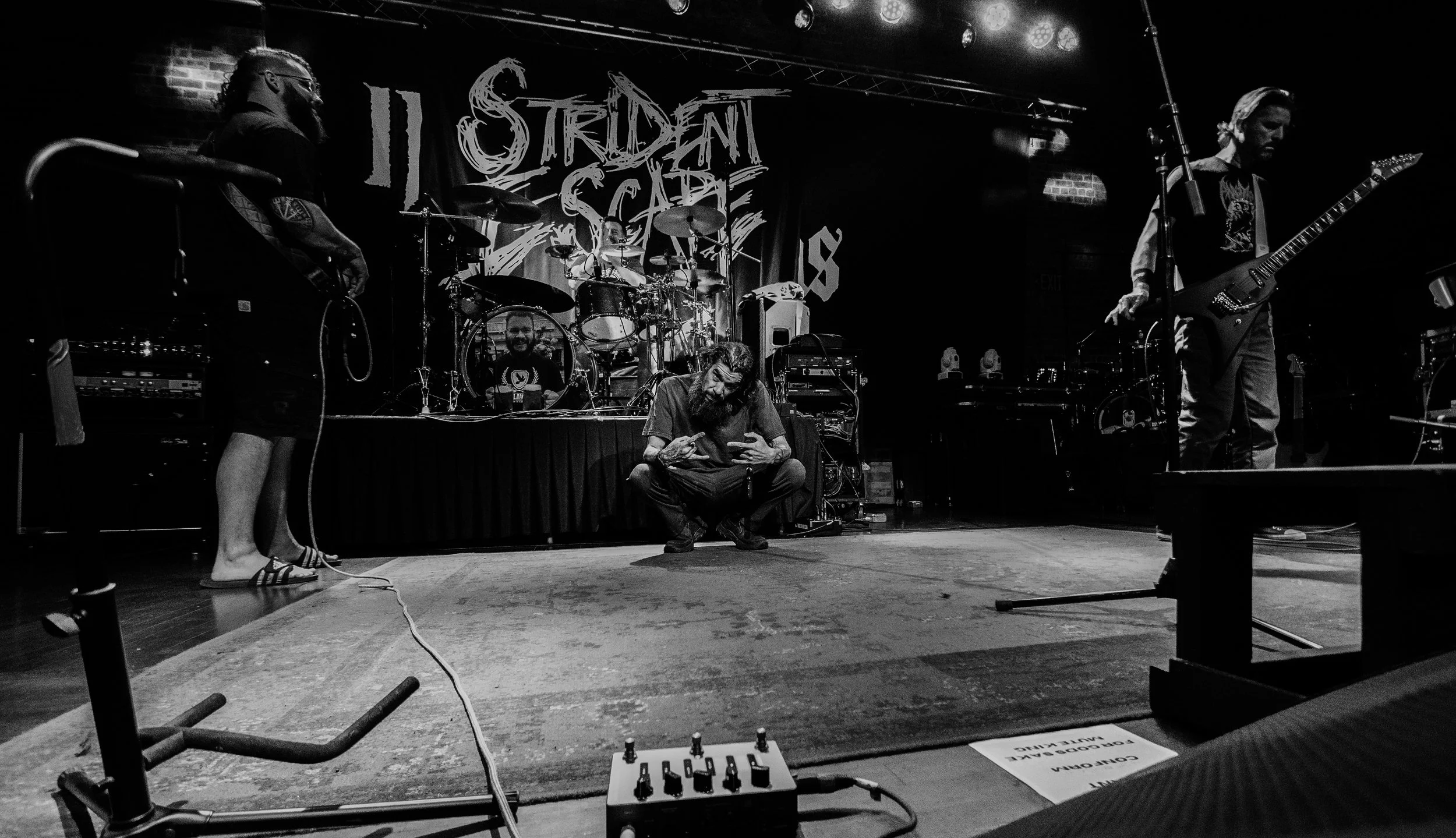 Black and white photo of a rock band performing on stage. Two guitarists, a drummer, and a person sitting on the floor looking at their phone. The backdrop has the band’s name, 'Lizard Stone,' displayed in a handwritten style.