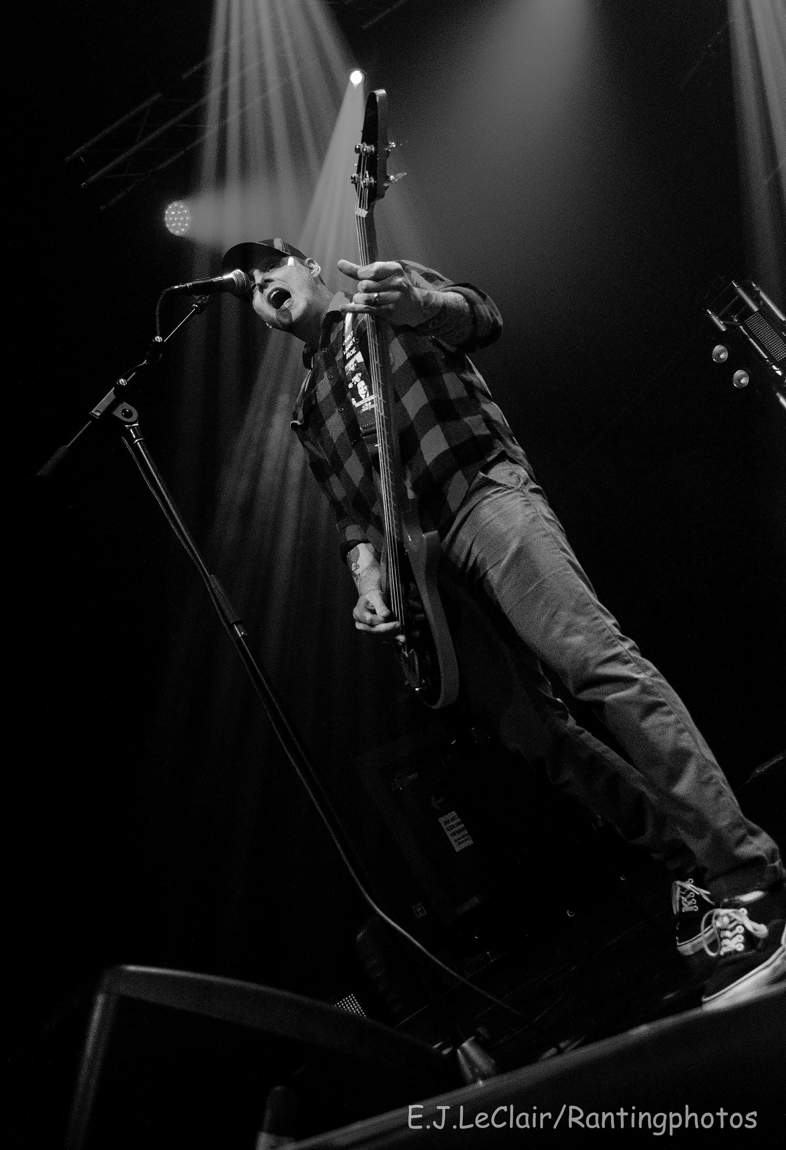 A black and white photo of a man singing into a microphone and playing an electric bass guitar on stage, with stage lights and equipment visible.