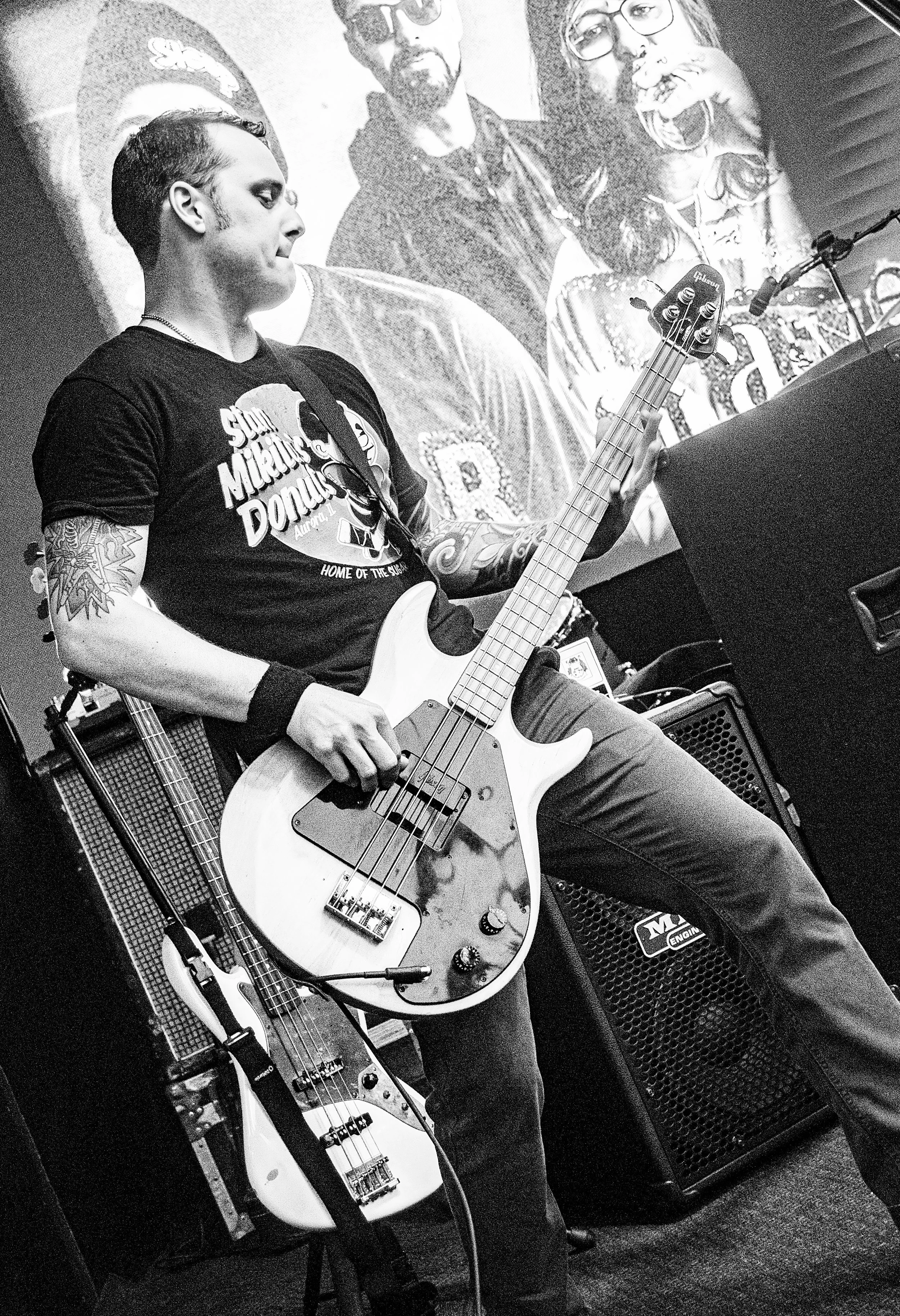 A person with tattoos on their arms playing an electric bass guitar on stage. They are wearing a T-shirt with the text 'Sun Microsystems Donuts' and a graphic of a donut with a bite taken out of it. In the background, there is a large poster or mural
