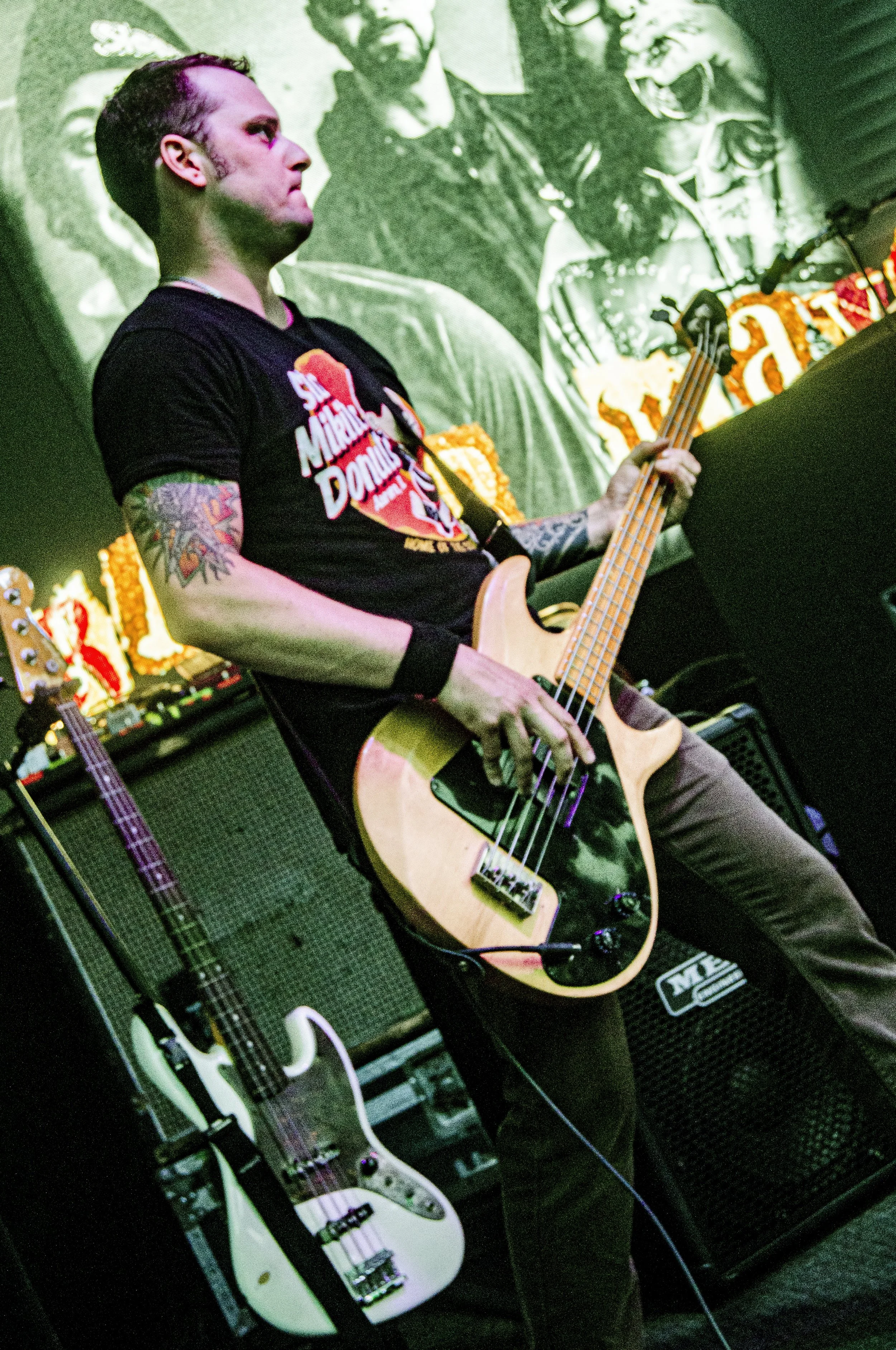 A man with tattoos playing a bass guitar on a stage with musical equipment and amplifiers, a large black-and-white poster of a group in the background.