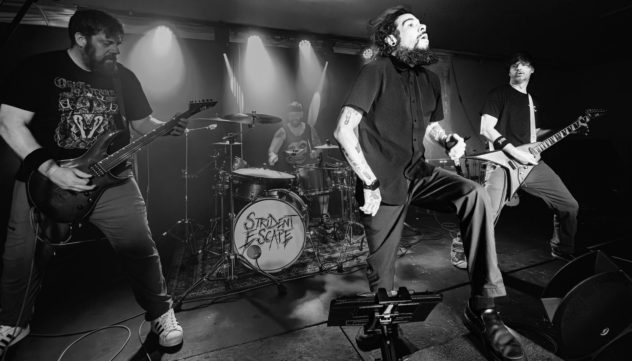 Black and white photo of a band performing live on stage. Four musicians are visible, with one singer holding a microphone. The band’s drum set has a logo that reads ‘Strident Escape’. The musicians are playing electric guitars and drums, with stage 