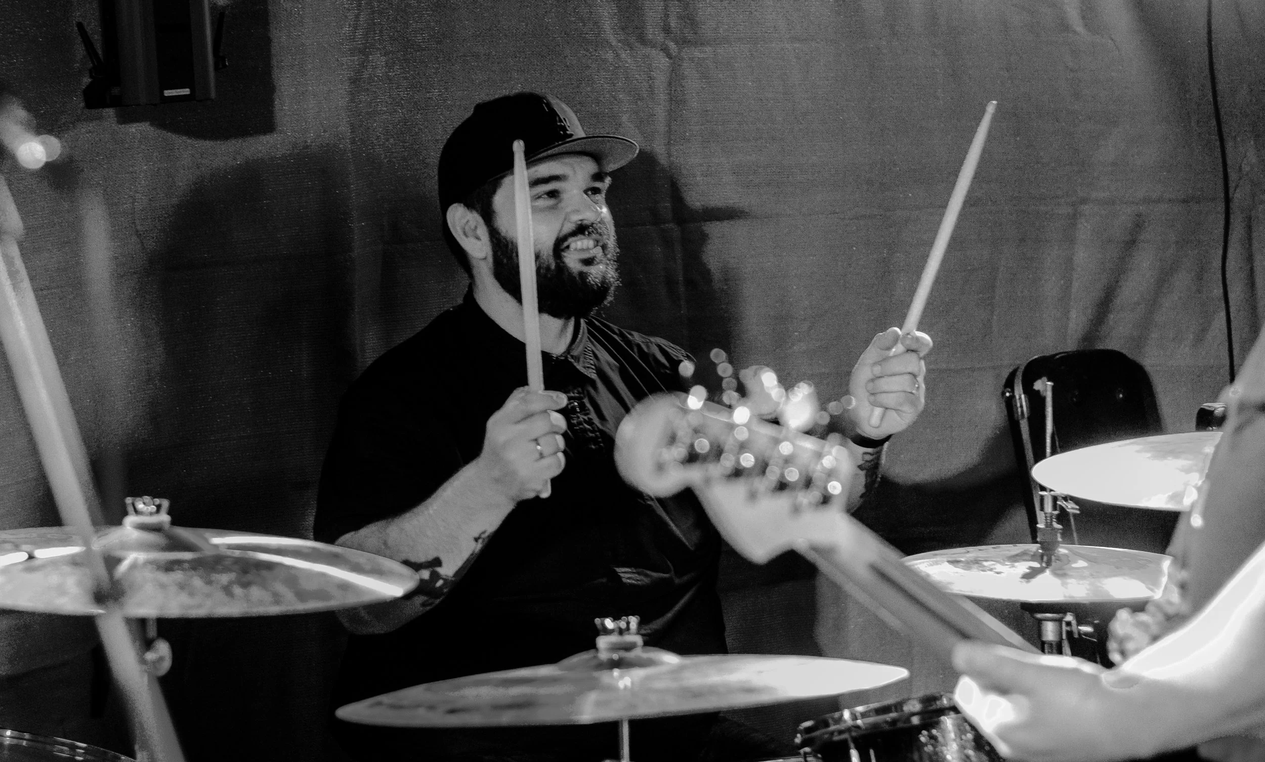 A man with a beard and a baseball cap playing the drums in a music studio, smiling while holding drumsticks.