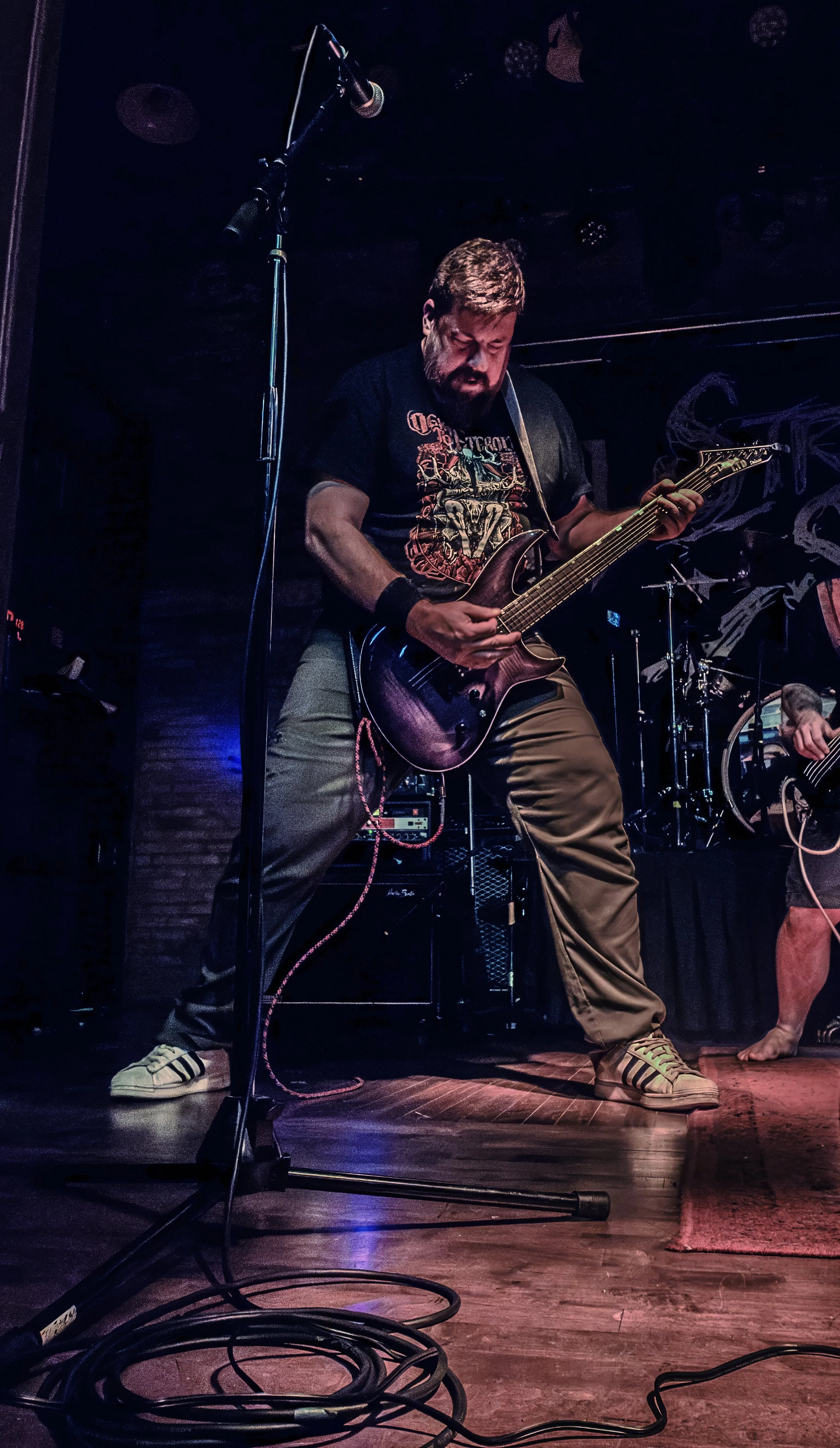 A man playing an electric guitar on stage during a performance, wearing a black graphic t-shirt, khaki pants, and striped sneakers, with stage equipment and a drum set visible in the background.