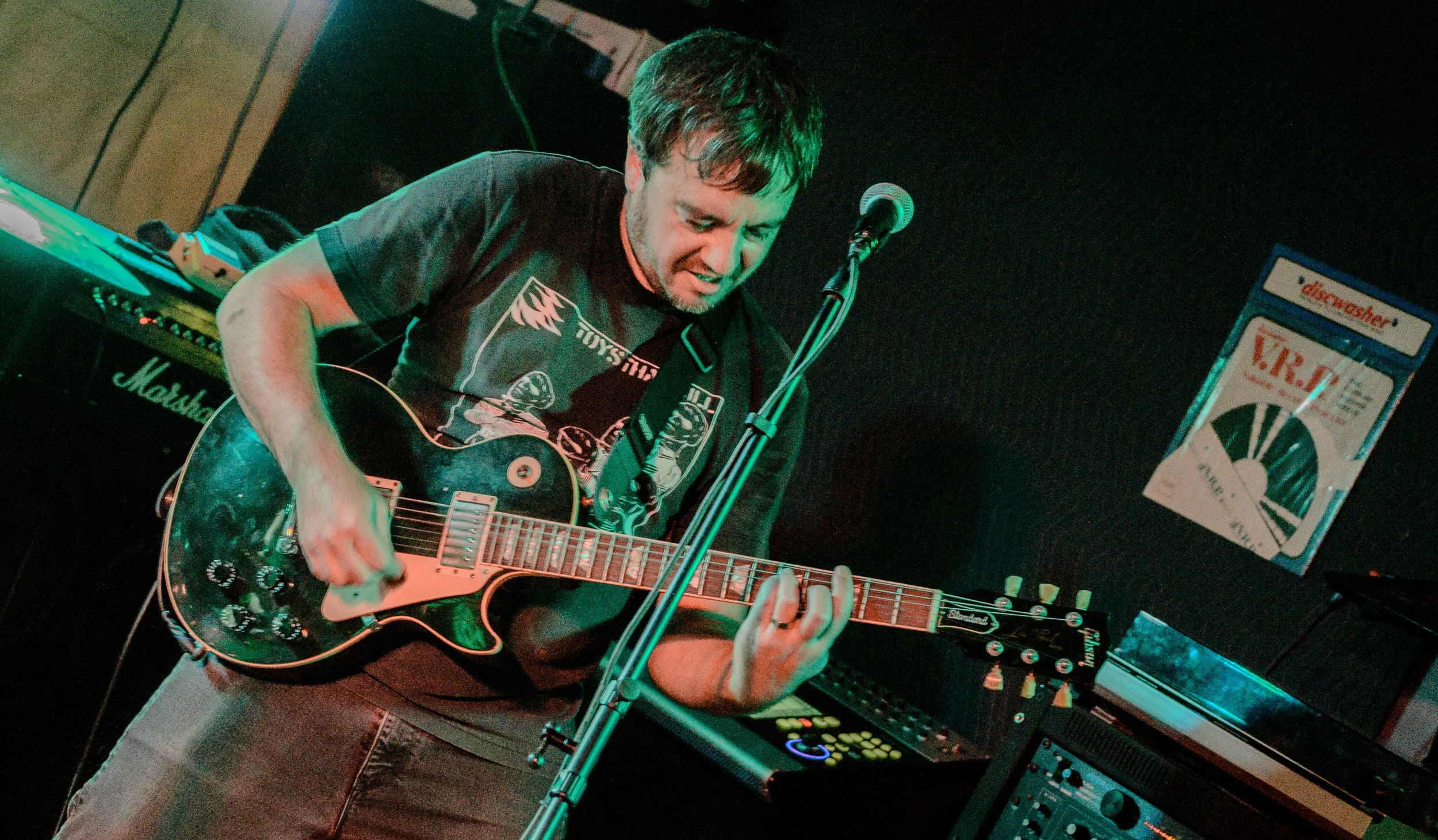 A man playing an electric guitar on stage with music equipment and a keyboard in the background, illuminated by green and red stage lights.