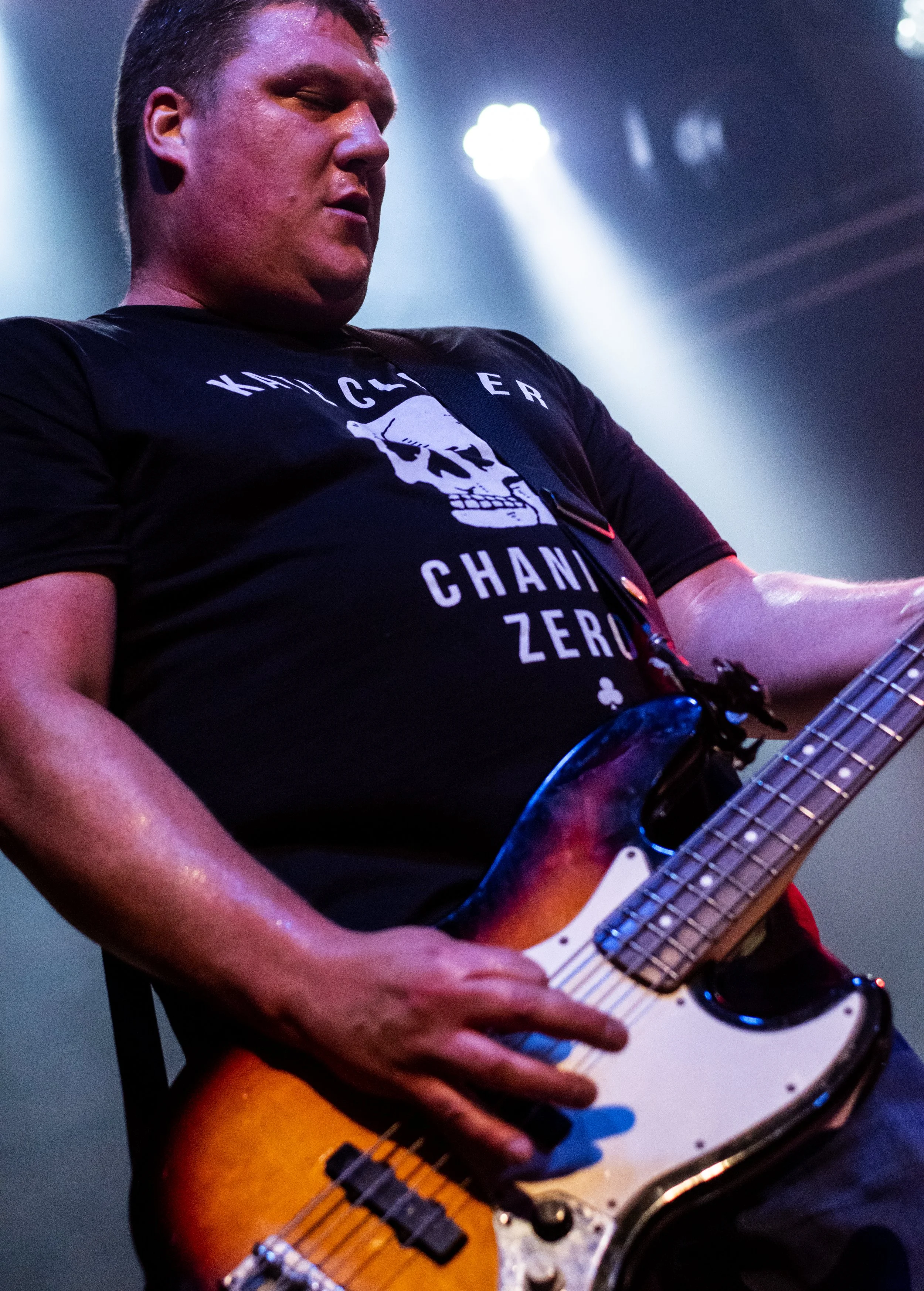 Close-up of a man playing an electric guitar on stage, wearing a black T-shirt with a skull graphic and text. The background shows stage lighting.