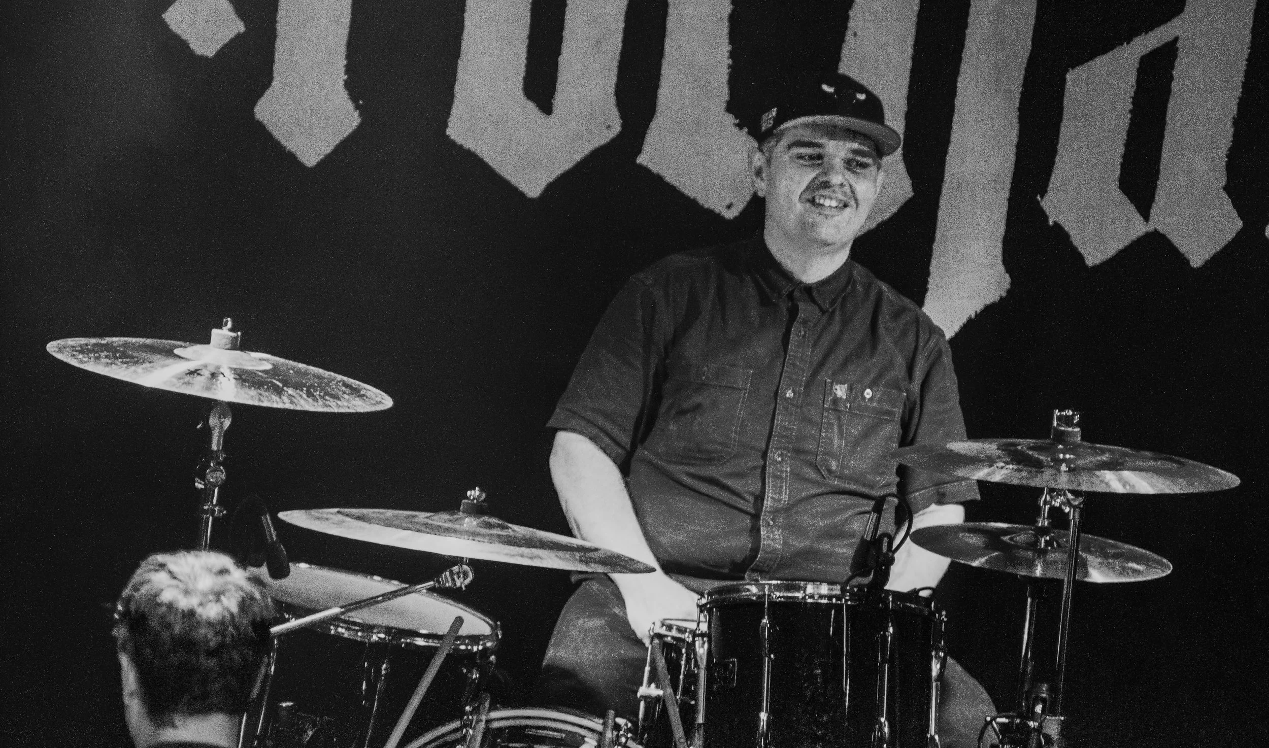 A young man wearing a cap and a short-sleeved shirt, smiling while sitting at a drum set, with a dark background featuring large, stylized letters.