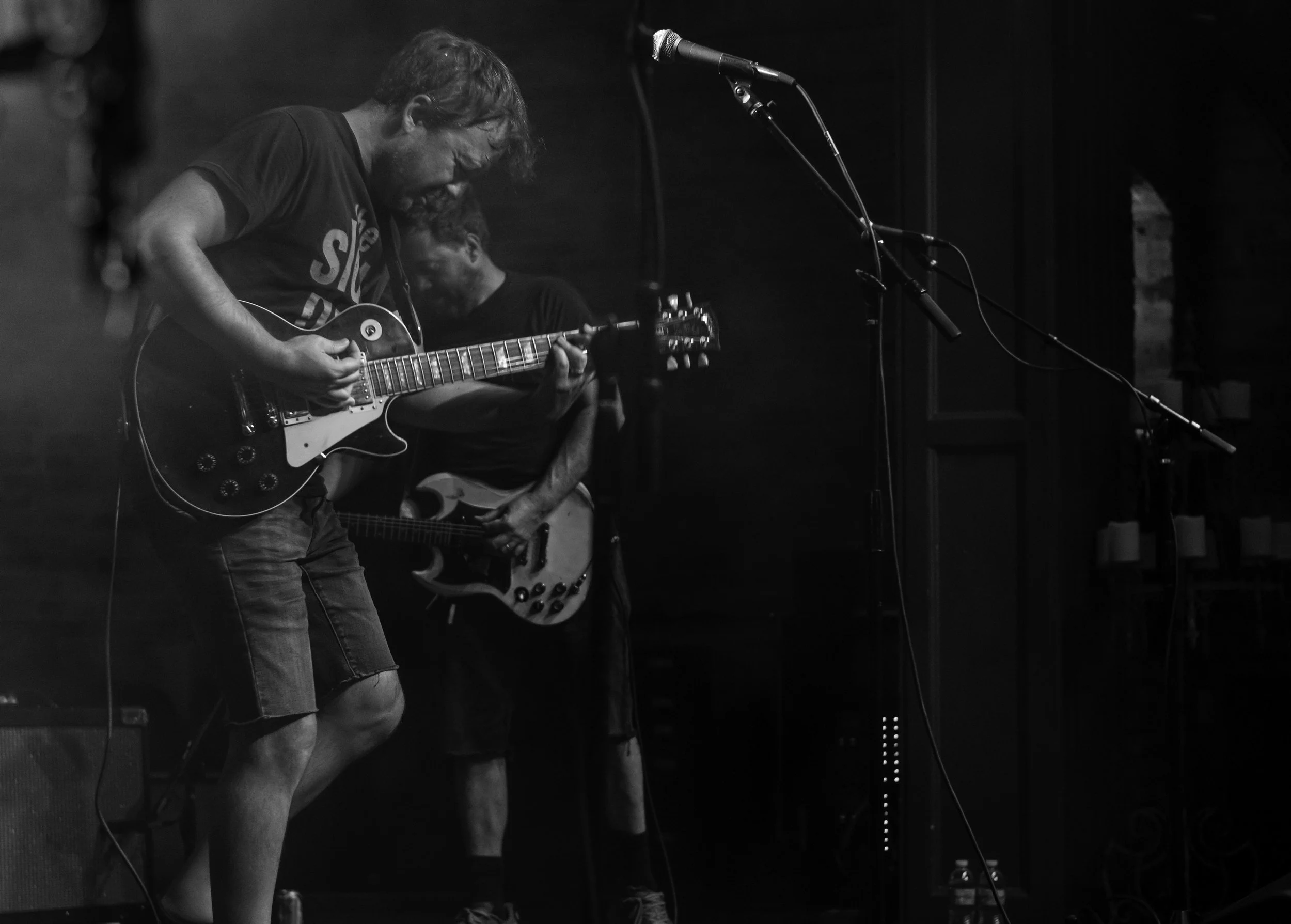 A black and white photo of a musician playing an electric guitar on stage, with a microphone in front of him, and another musician playing a bass guitar in the background.