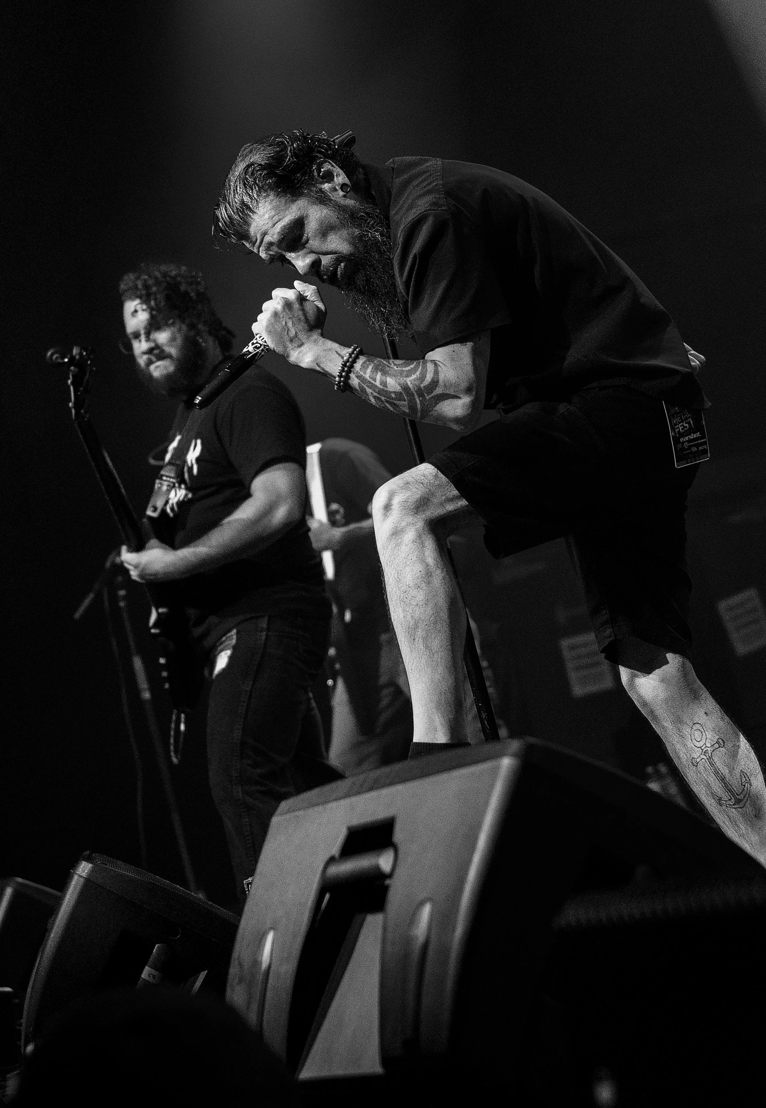 A black-and-white photo of a man with a beard and tattoos, holding a microphone with his eyes closed, leaning forward, in a music performance setting.