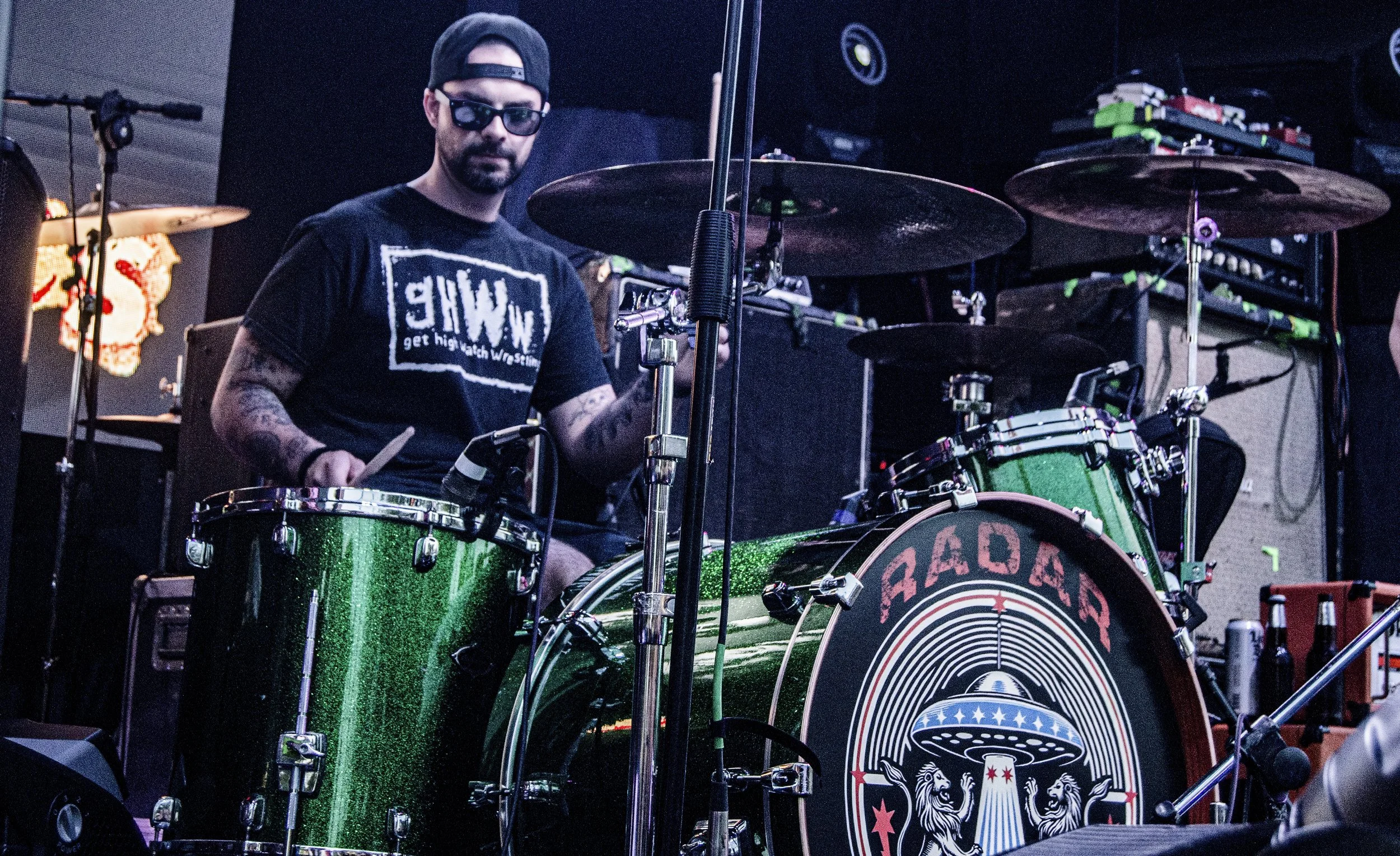 A man wearing a black cap, sunglasses, and a black T-shirt is sitting behind a green glittery drum set, holding drumsticks. The drum set has a bass drum with a logo that appears to have the name 'RABiC' and features a design with a lion and stars. Th