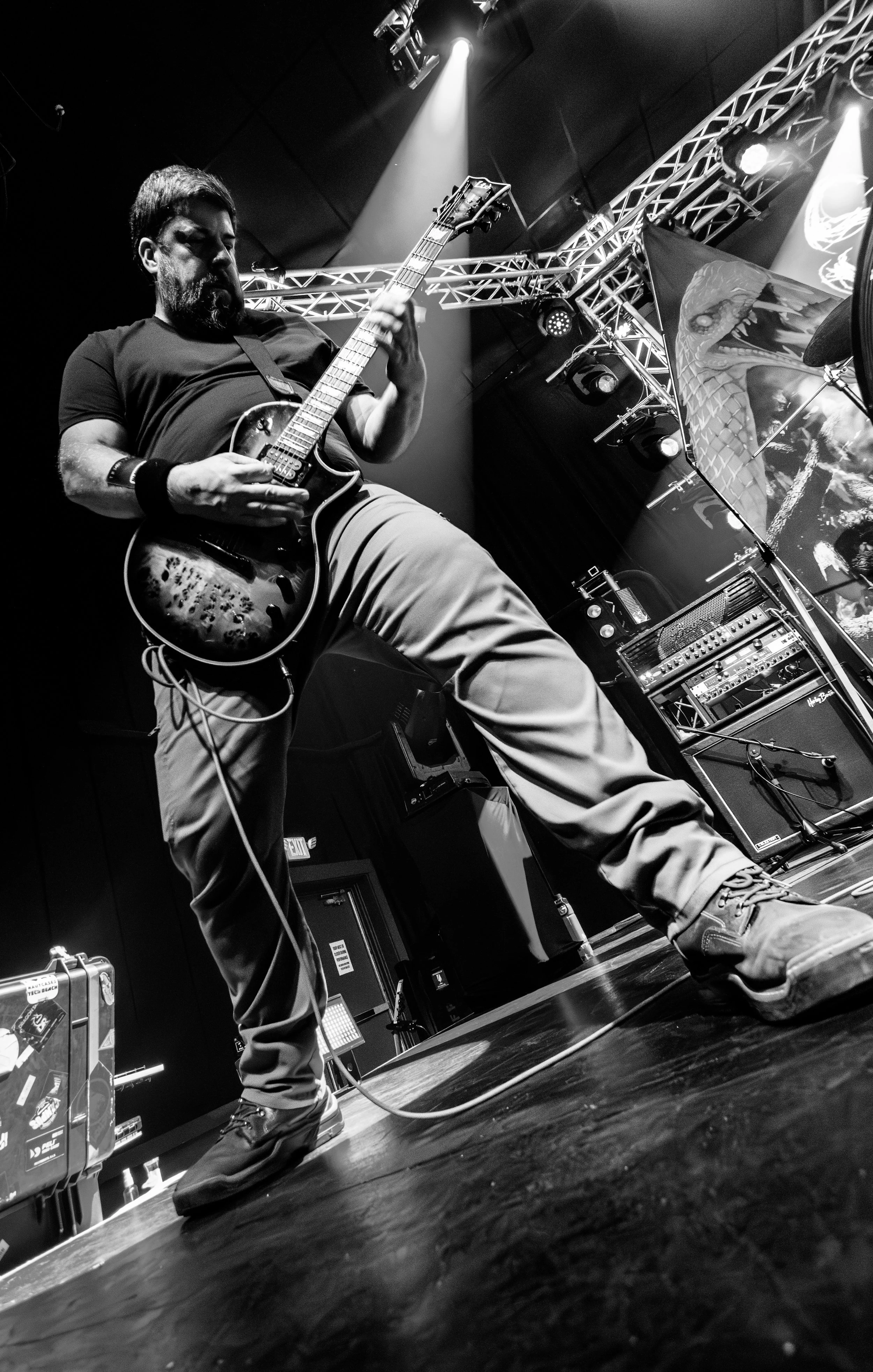 A musician playing an electric guitar on stage, shot from a low angle in black and white with stage lights and equipment in the background.