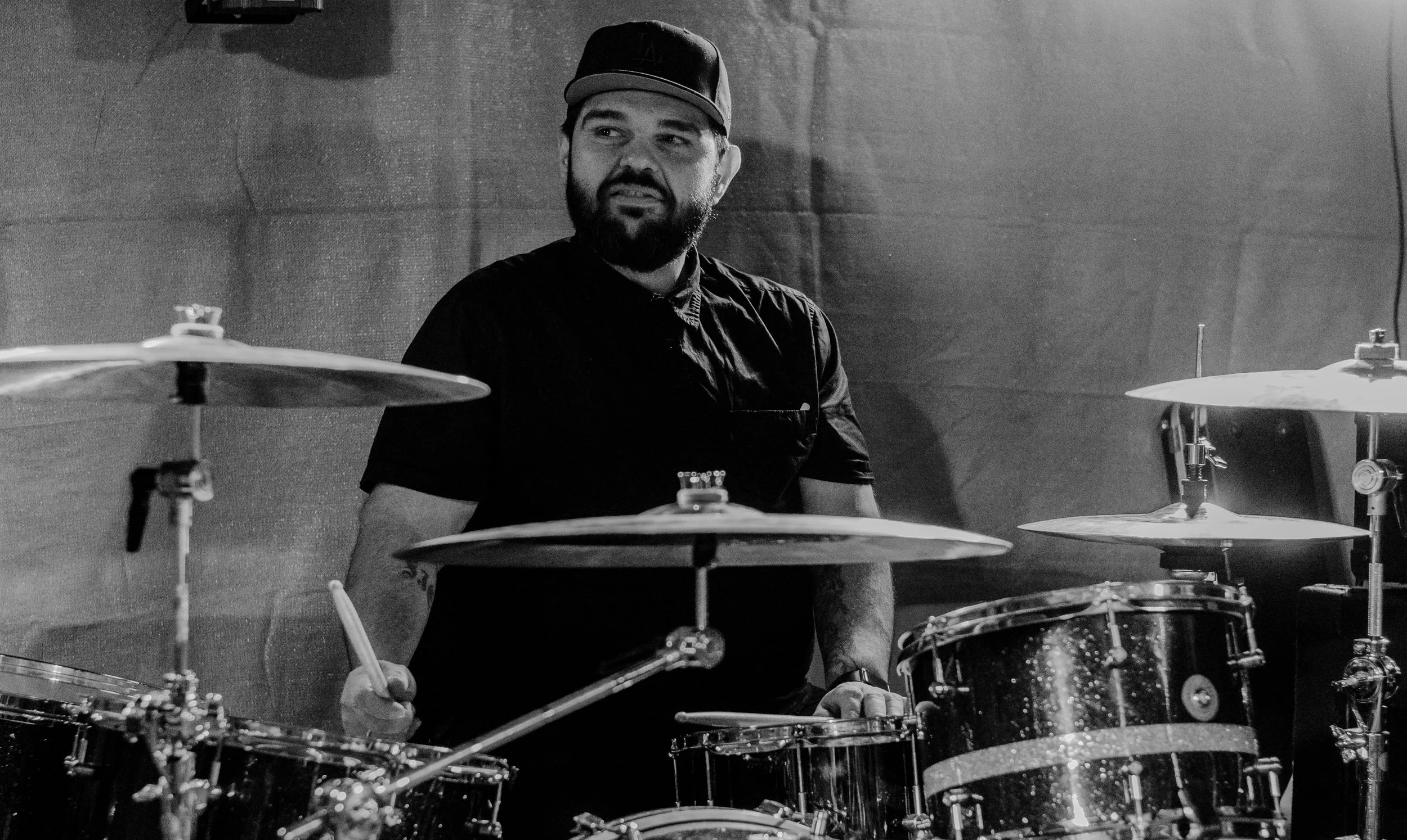 A man with a beard wearing a baseball cap and a black shirt playing the drums in a music studio.