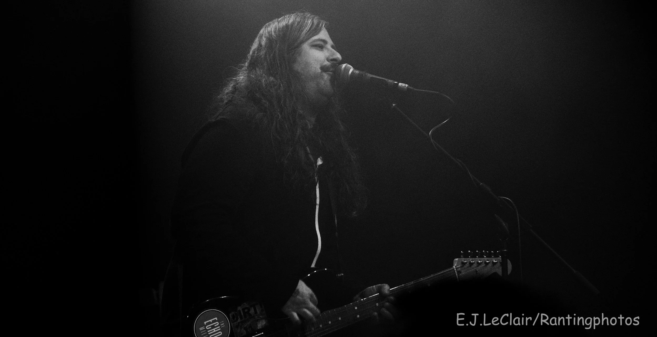 Black and white photo of a person with long hair playing an electric guitar and singing into a microphone on stage.