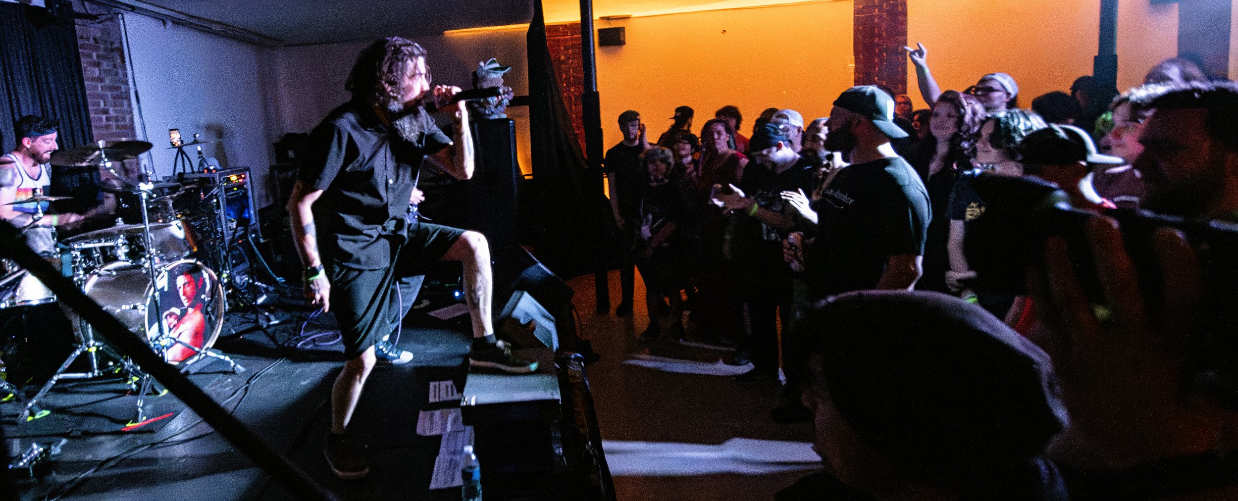 A band performs live on stage at a concert with a lead singer, drummer, and audience dancing.
