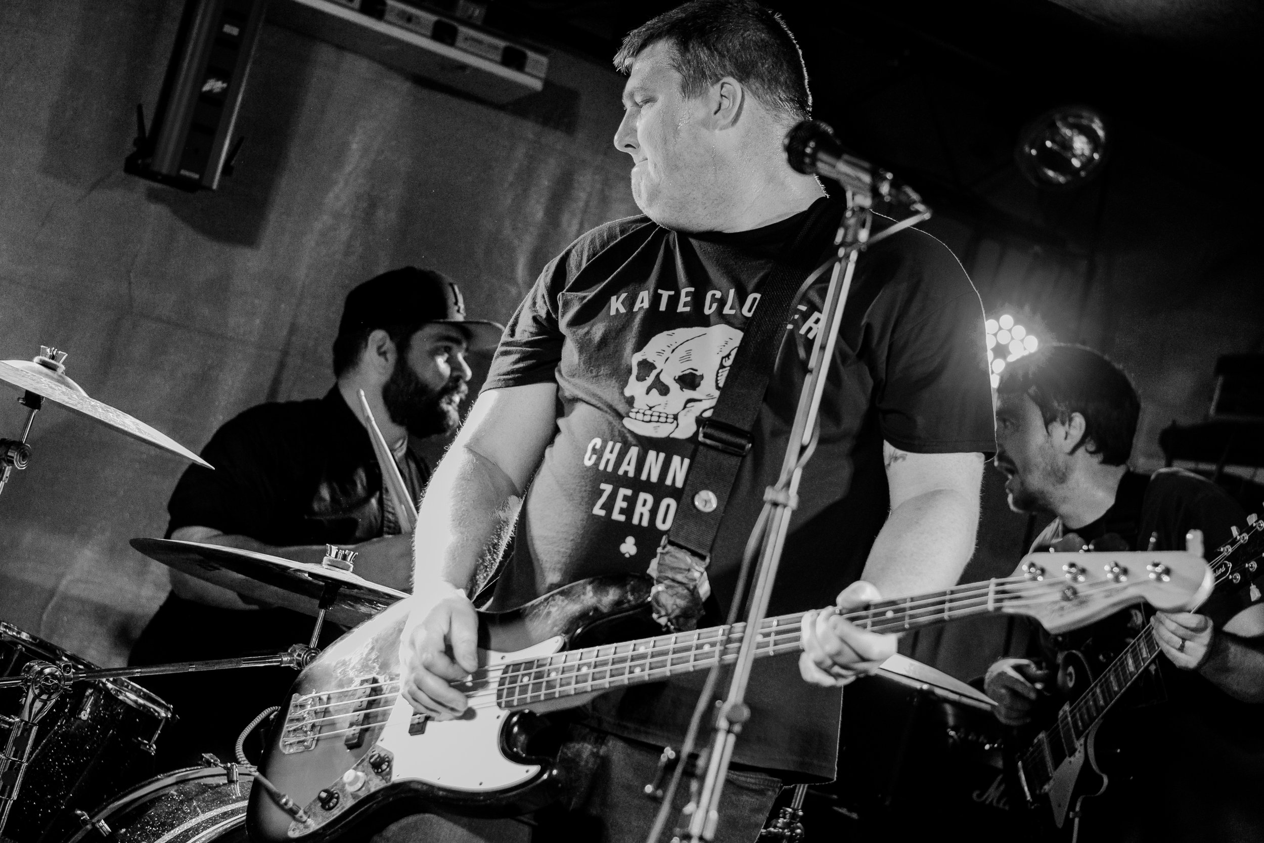 Three musicians performing on stage: a guitarist in the foreground with a bass guitar, a drummer in the background, and a second guitarist on the right. The scene is in black and white.