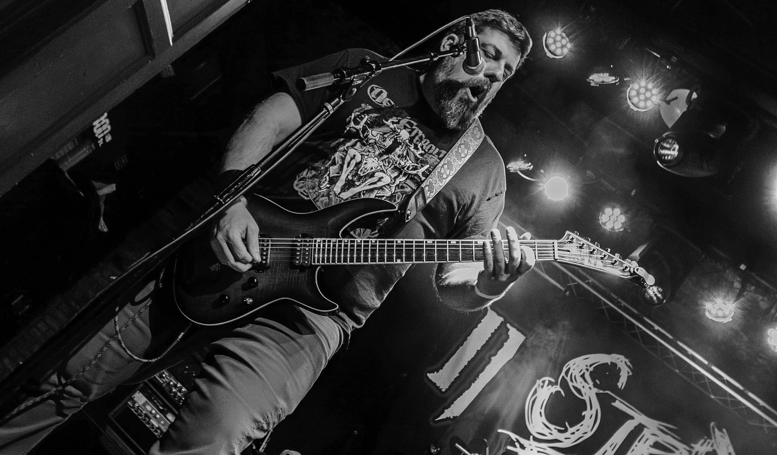 A male musician with a beard and mustache playing an electric guitar and singing into a microphone during a live performance at a dark venue with stage lights.