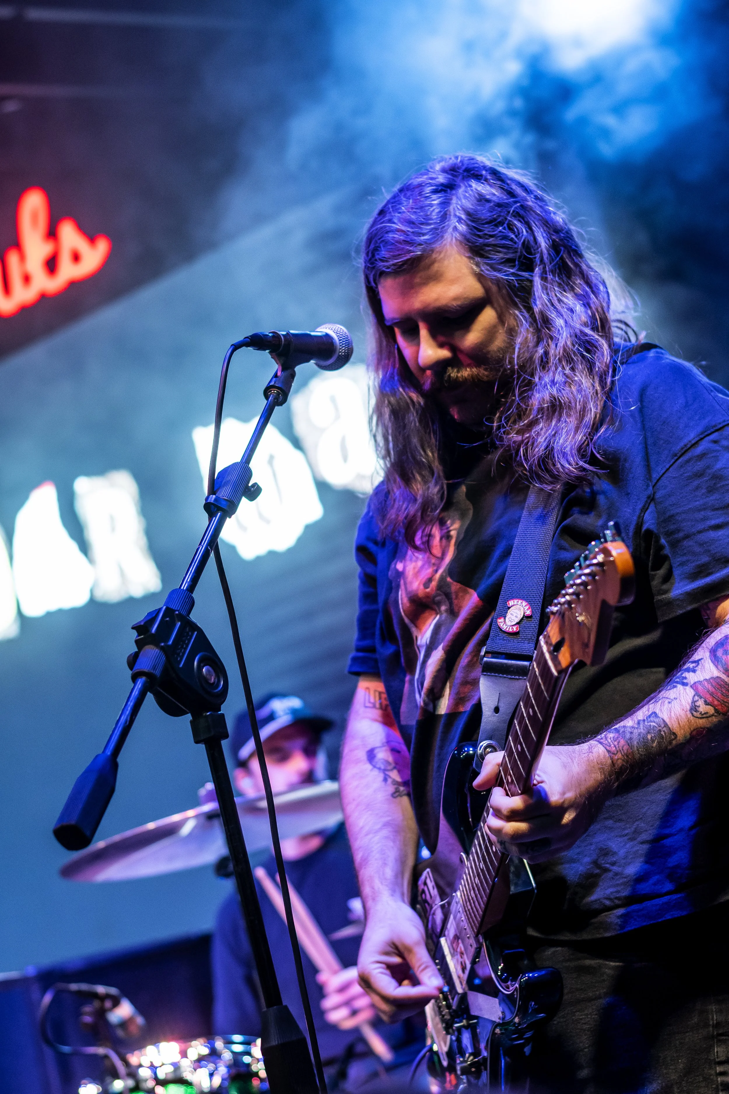 Musician with long hair and tattoos playing an electric guitar on stage with a microphone and drums in the background, illuminated by blue and purple stage lighting.