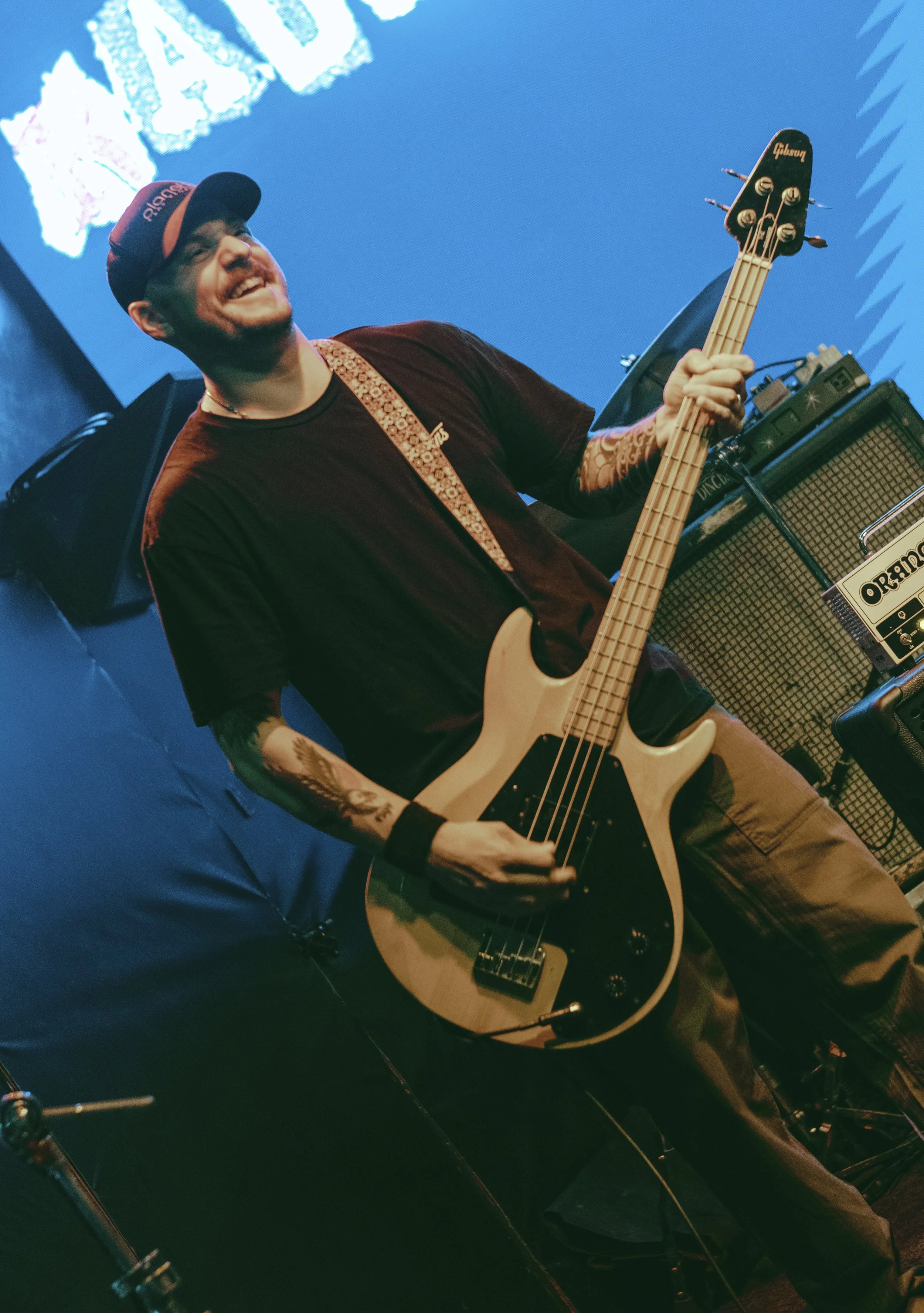 A smiling man with a beard and glasses playing an electric guitar on a stage with a blue background and large screen display.