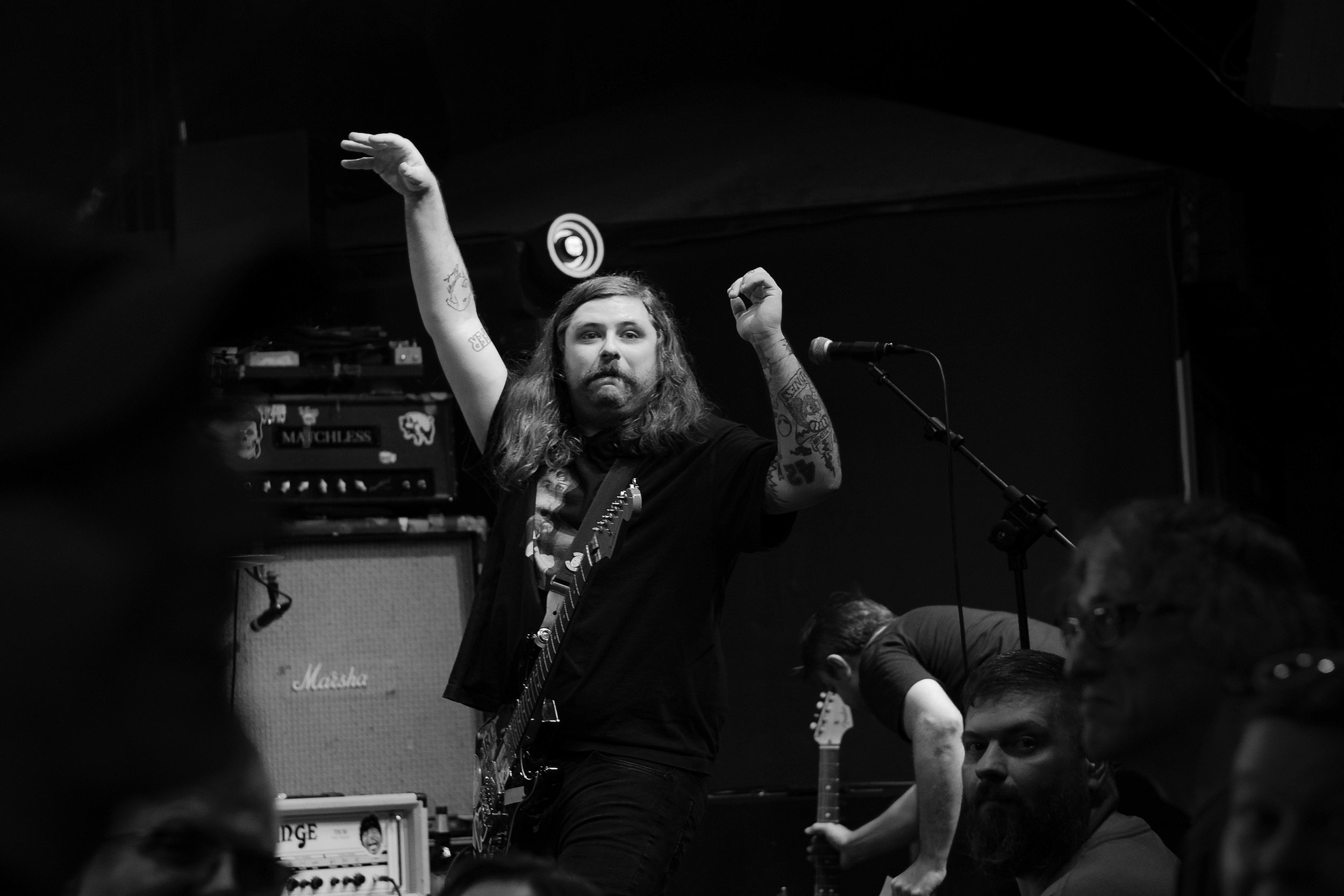 A musician with long hair playing an electric guitar during a live performance with his hand raised, surrounded by musical equipment and a dark background.