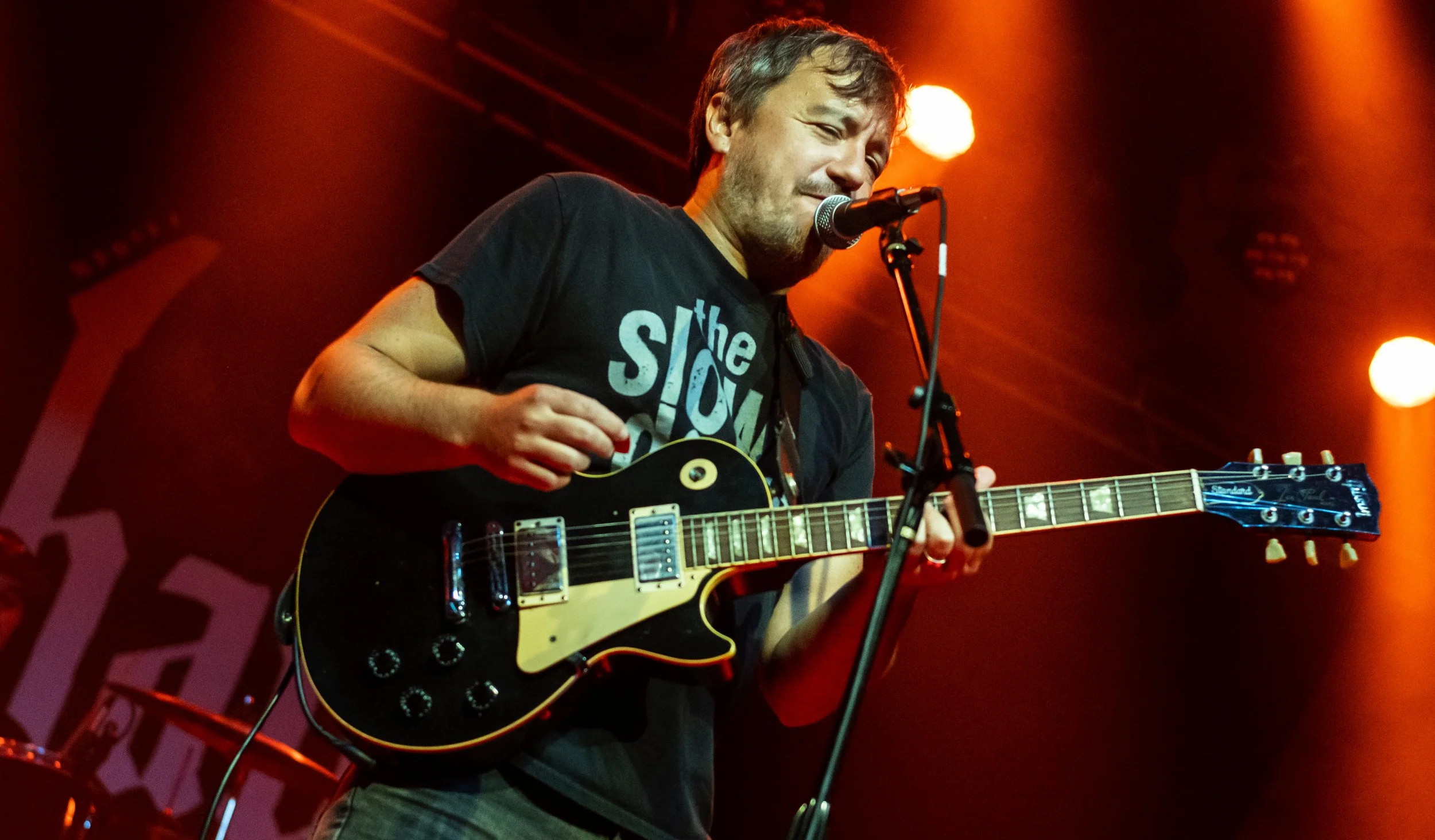 A man performing on stage with an electric guitar, singing into a microphone, illuminated by warm orange stage lights.