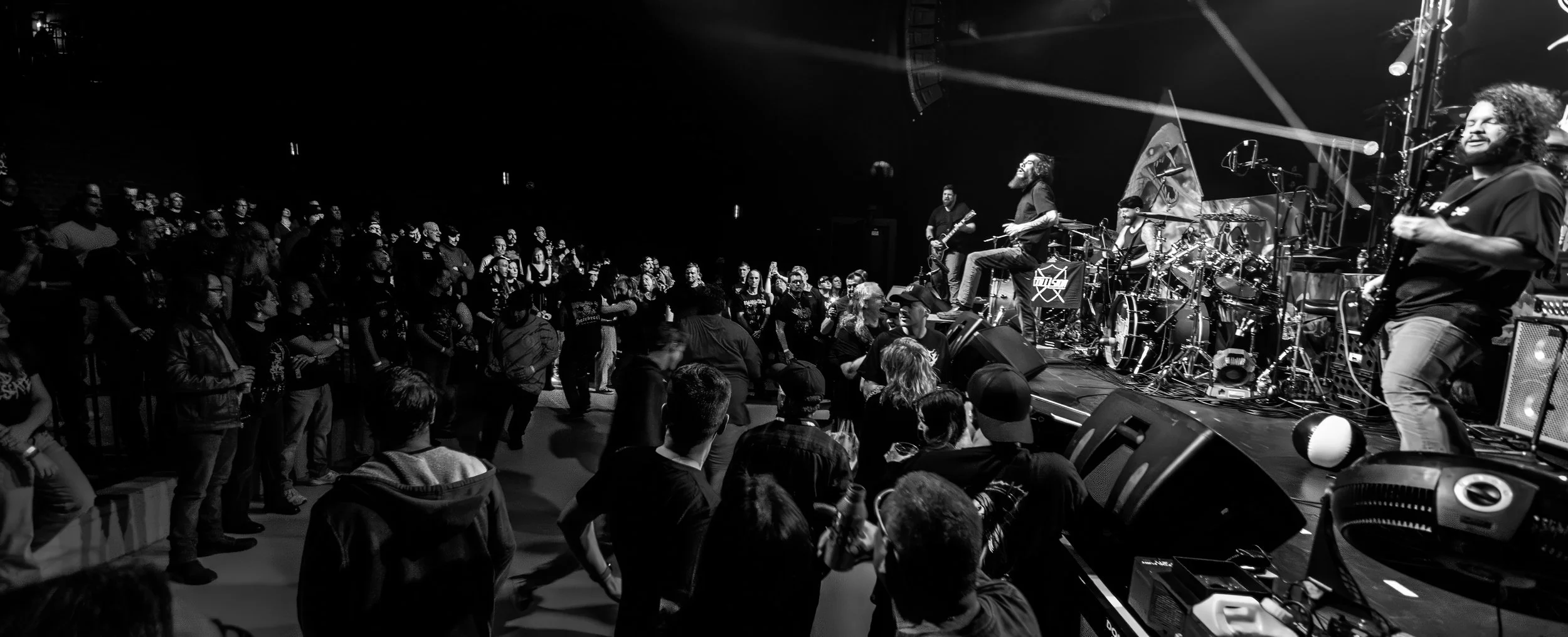 Black and white photo of a live concert with a band performing on a stage and an audience watching, some standing and some seated.