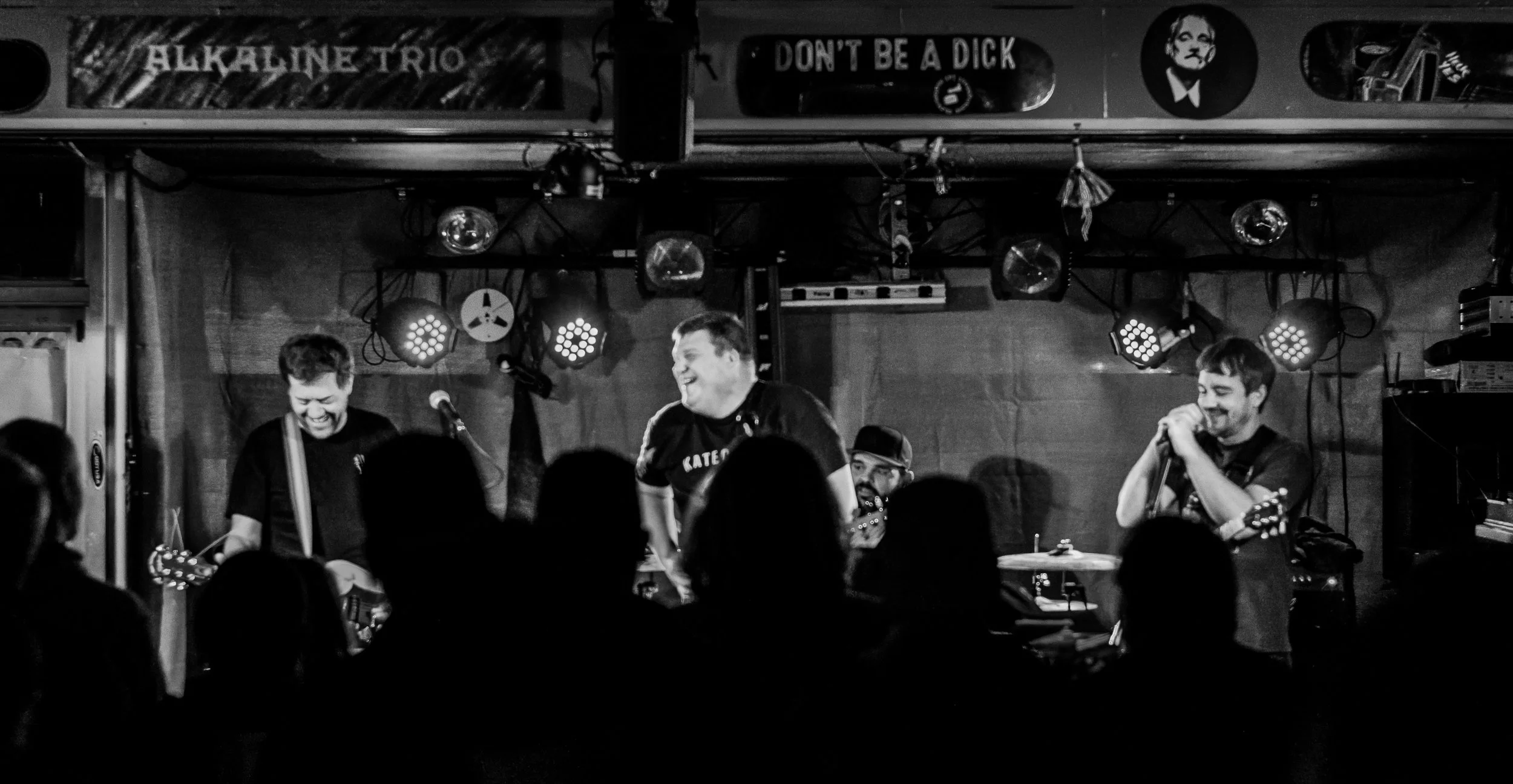 Black and white photo of a band performing on a stage with three members smiling and playing instruments, audience in foreground, stage with lighting and signs overhead.