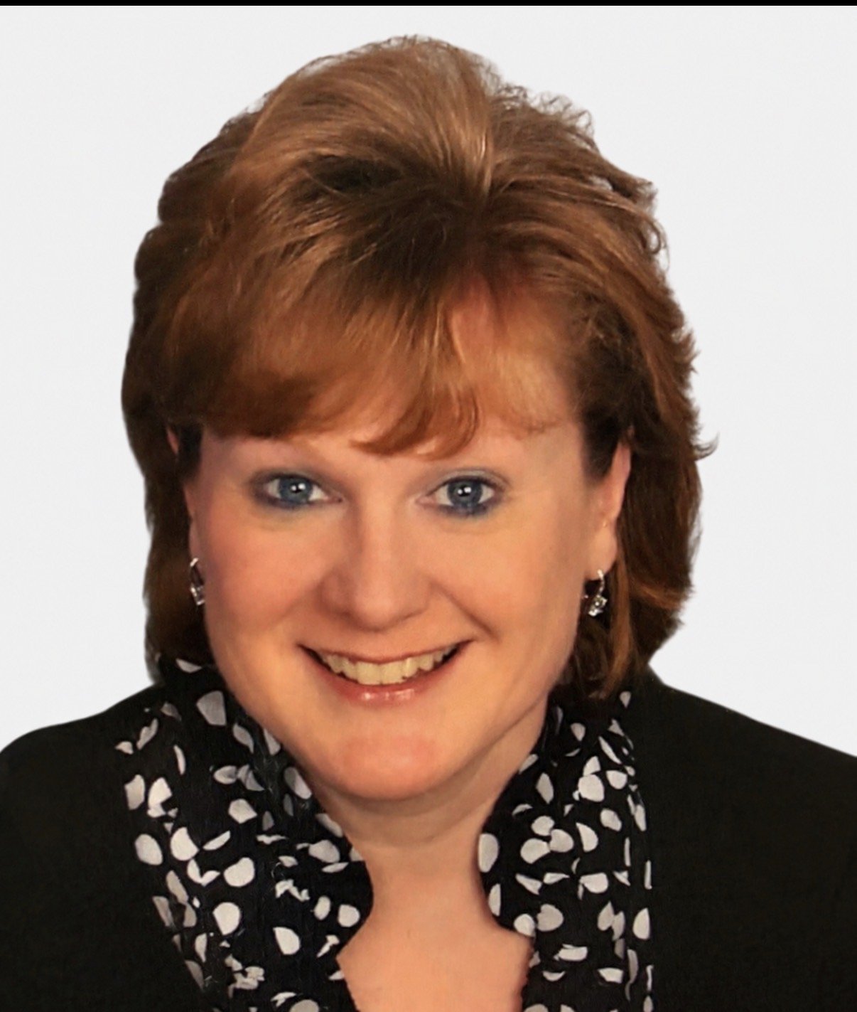 A woman with short, wavy, reddish-brown hair and blue eyes, smiling, wearing earrings, a black jacket, and a black and white patterned blouse against a plain light background.