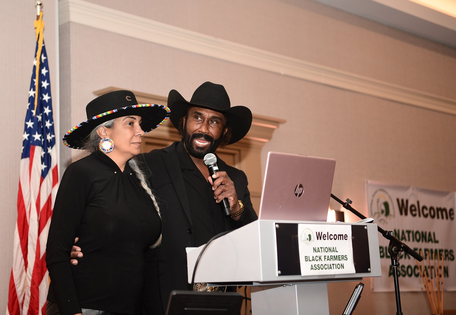 A man and woman wear cowboy hats and stand at a podium with a microphone. The woman has long gray hair, large earrings, and black clothing. The man has a beard, wears a black outfit, and is speaking into the microphone. An American flag and a banner for the National Black Farmers Association are visible in the background.