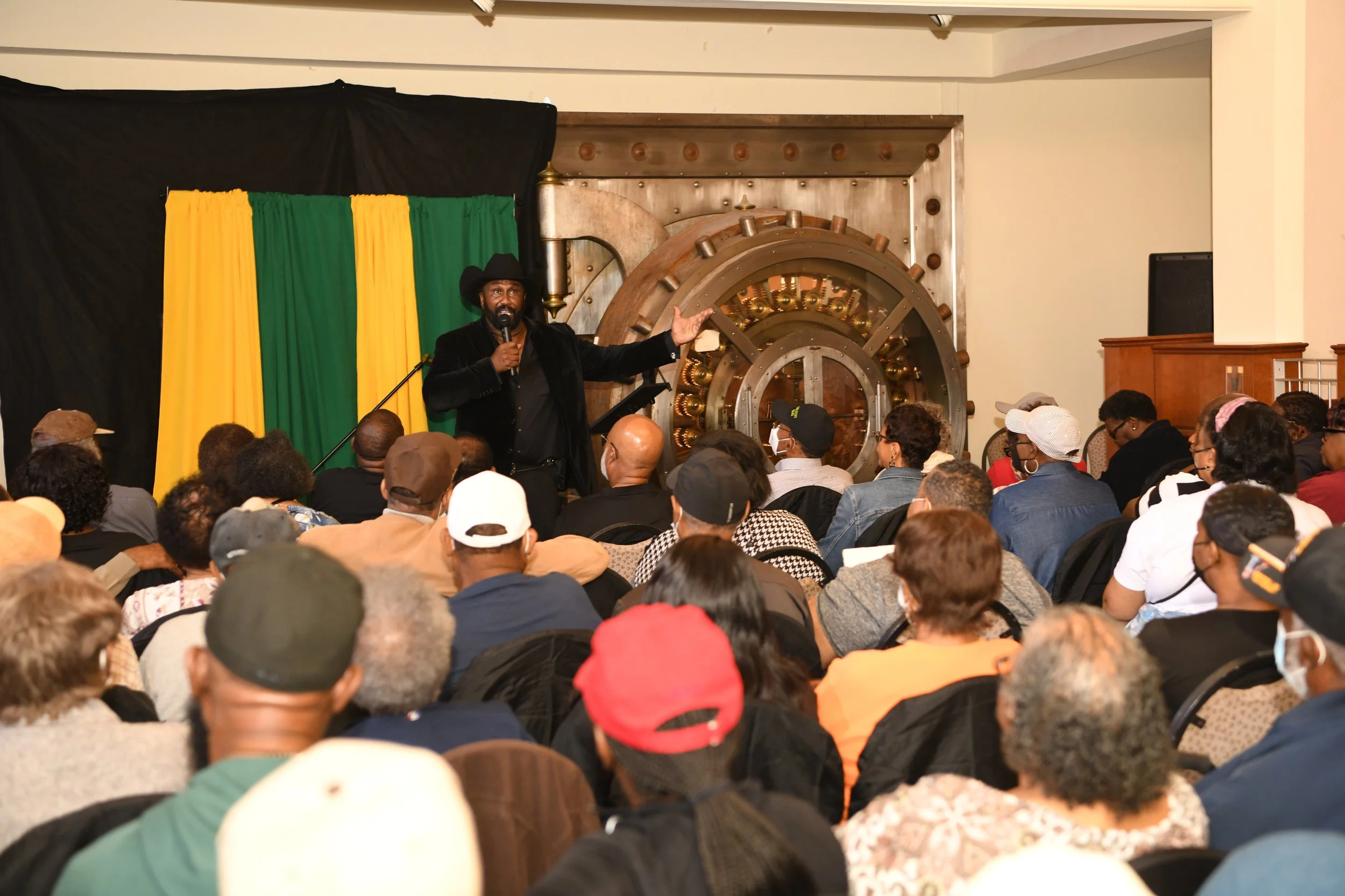 A man with a beard, wearing a black hat and black jacket, is speaking into a microphone while gesturing with his right hand. He is standing in front of a crowd at an event, with a background featuring black, yellow, and green curtains and a large, round, metallic, vault-like door.