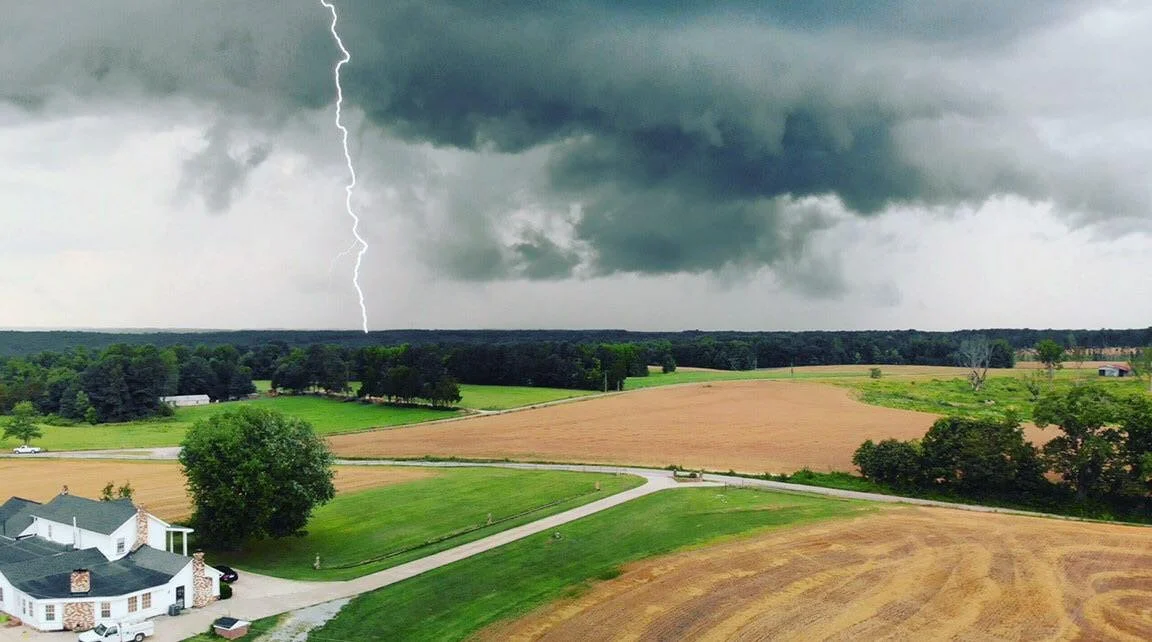 A rural landscape with storm clouds and a lightning bolt striking in the distance over open fields and a few houses.
