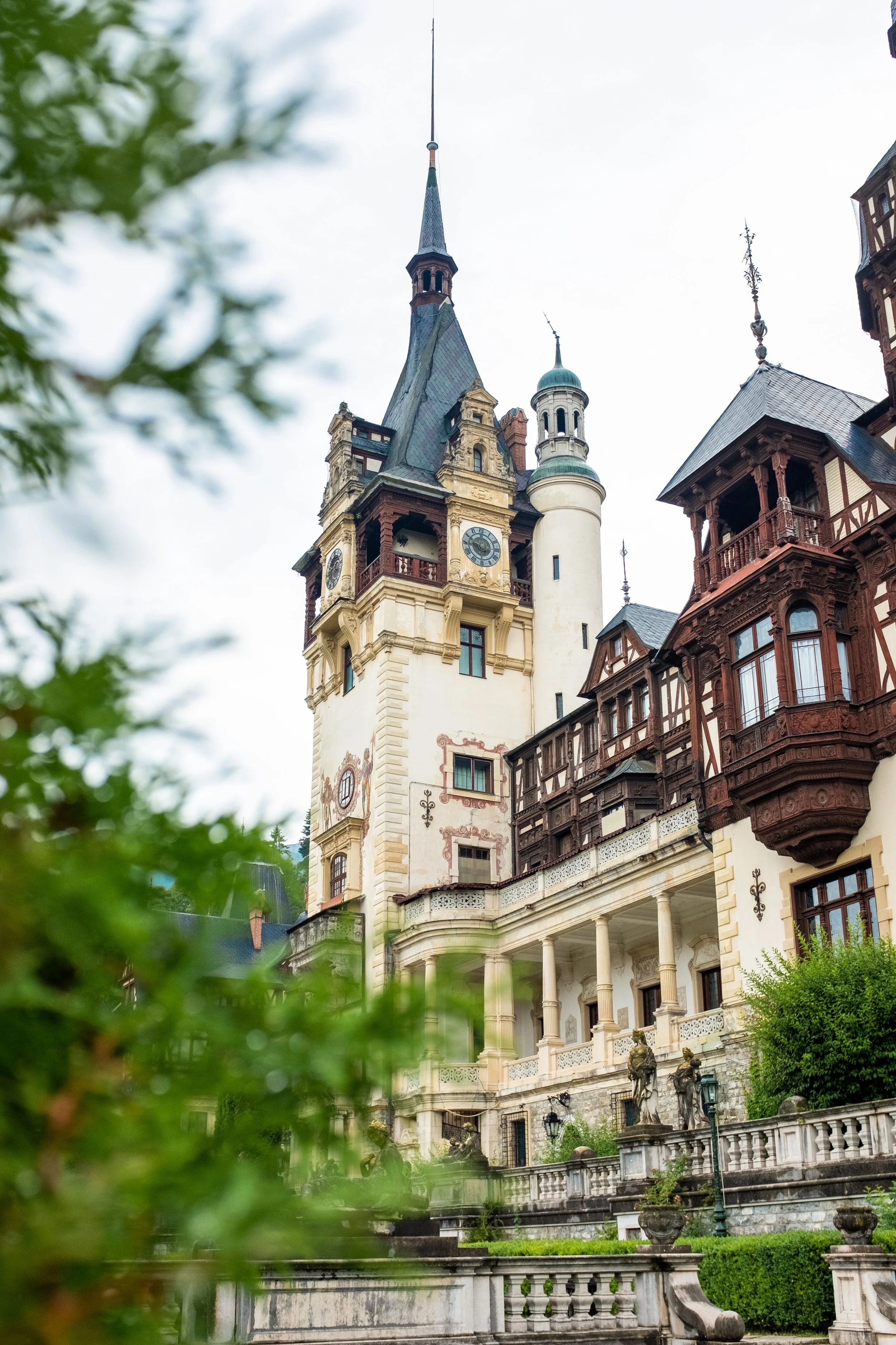 A grand historic building with a clock tower and turrets, featuring ornate architectural details, situated behind lush greenery and stone balustrades.