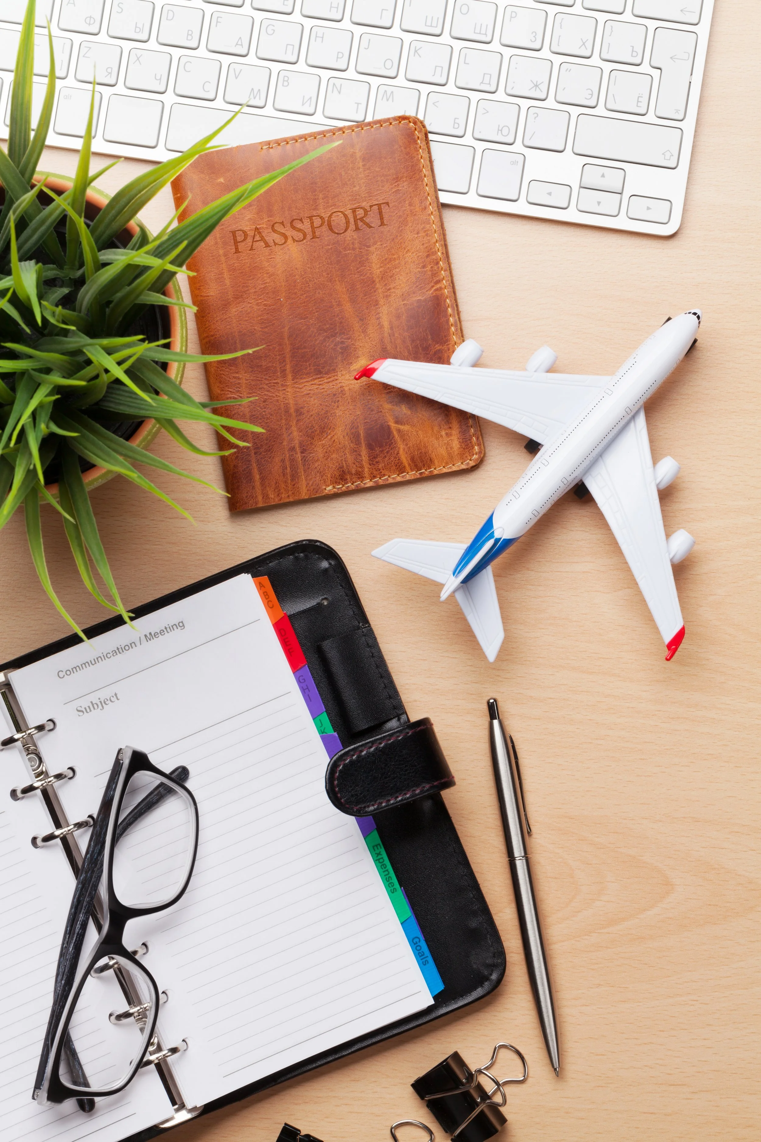 A desk with a potted plant, a leather passport holder, a white computer keyboard, a toy airplane, a planner with colorful tabs, a pair of black glasses, a silver pen, and various office supplies such as binder clips and a pen.