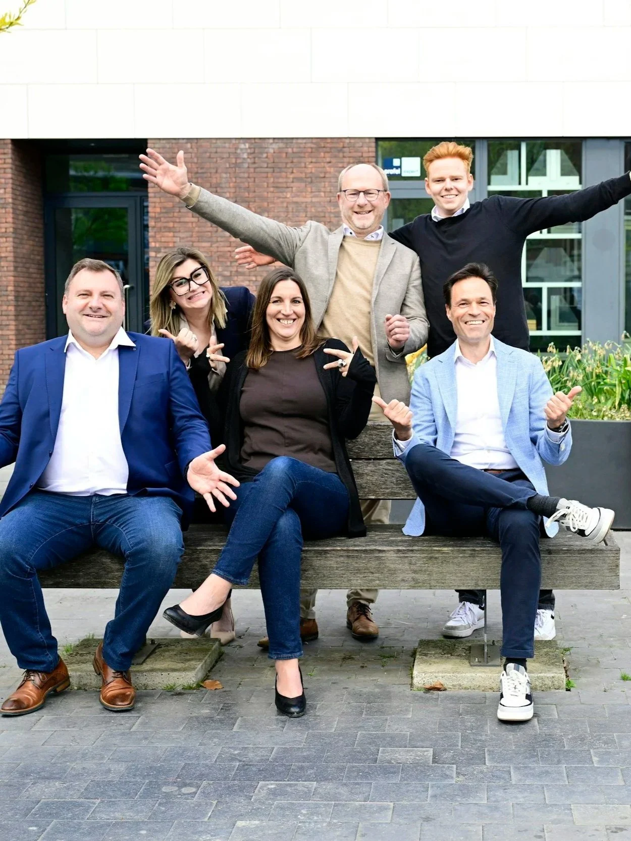 Group of six diverse adults sitting on a wooden bench outdoors, smiling and making animated gestures, with modern building background and greenery.