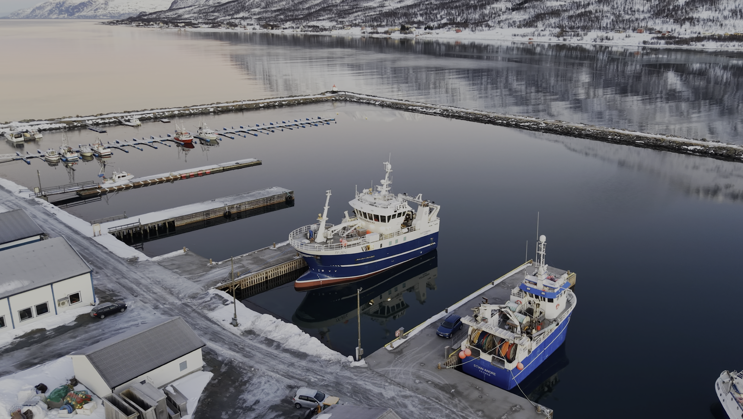 An aerial view of a snowy harbor with several boats docked, including a large blue and white ship. Surrounding the harbor are snow-covered buildings, roads, and mountainous terrain in the background reflecting on the calm water.