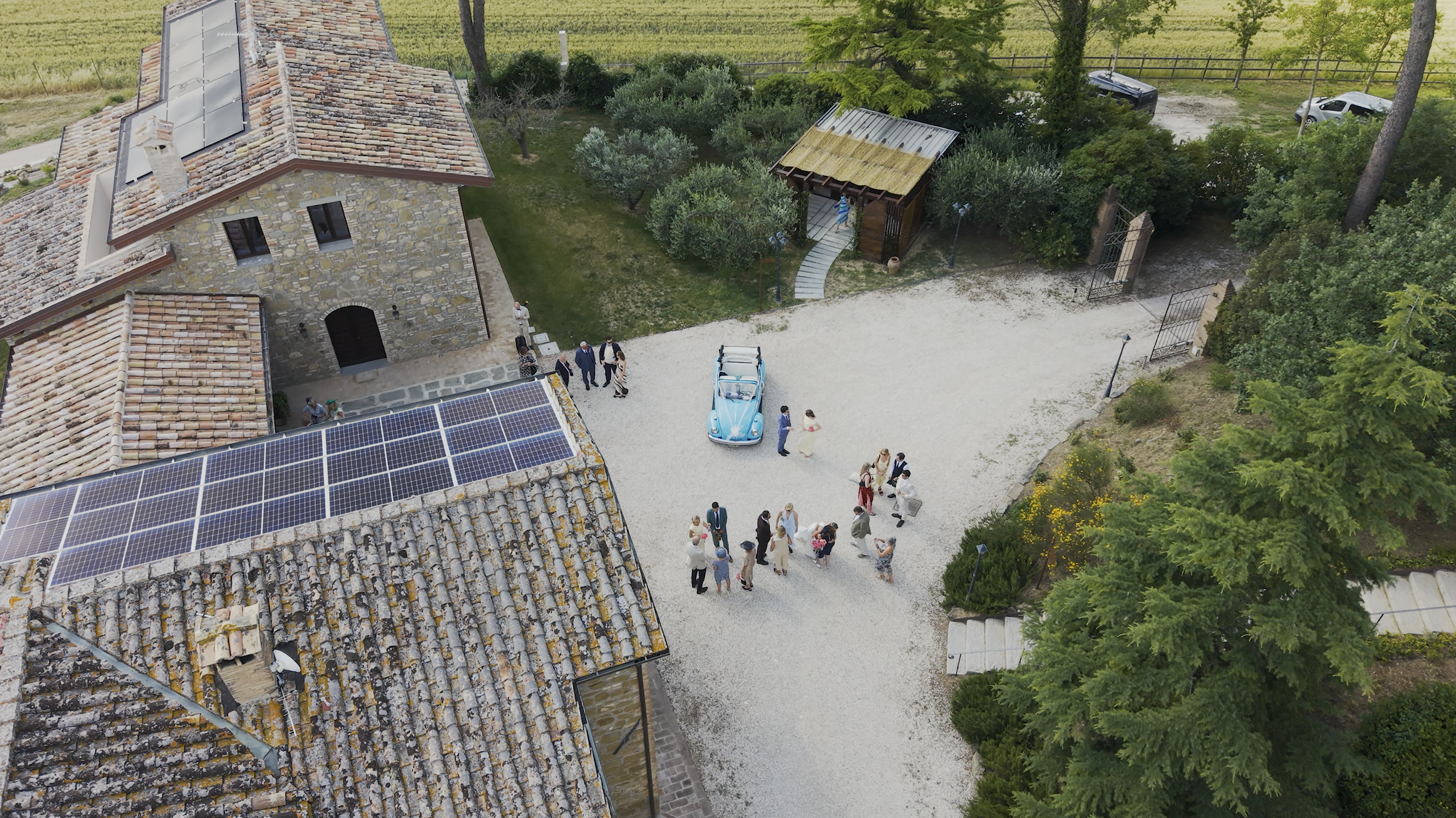 Aerial view of a rustic stone building with solar panels on the roof, surrounded by trees and greenery. There is a gravel courtyard with a vintage blue car, and a group of people gathered, indicating a social event or gathering.