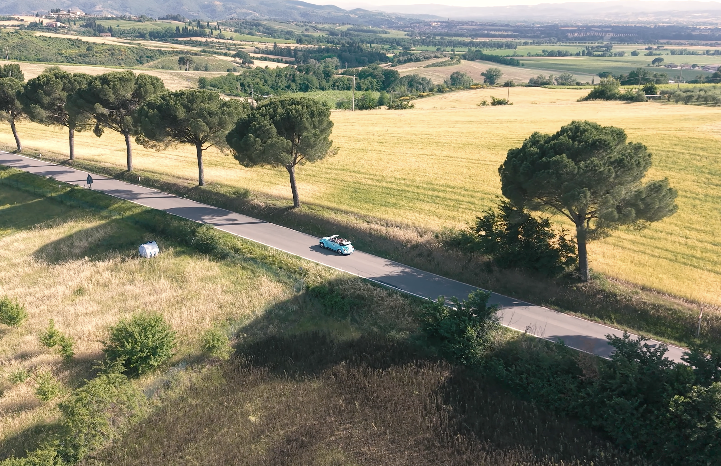 An aerial view of a rural landscape with rolling yellow fields, green trees, a paved road with a small vintage car, and distant hills under a partly cloudy sky.