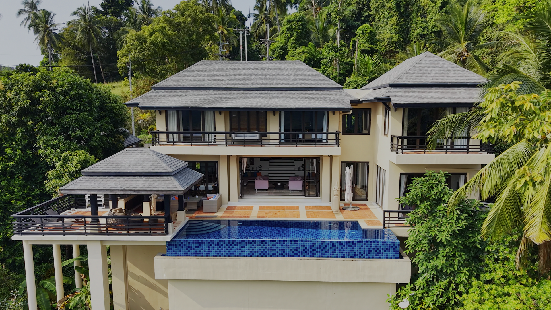 Large modern house with multiple balconies, outdoor patio, swimming pool, surrounded by lush green tropical trees.
