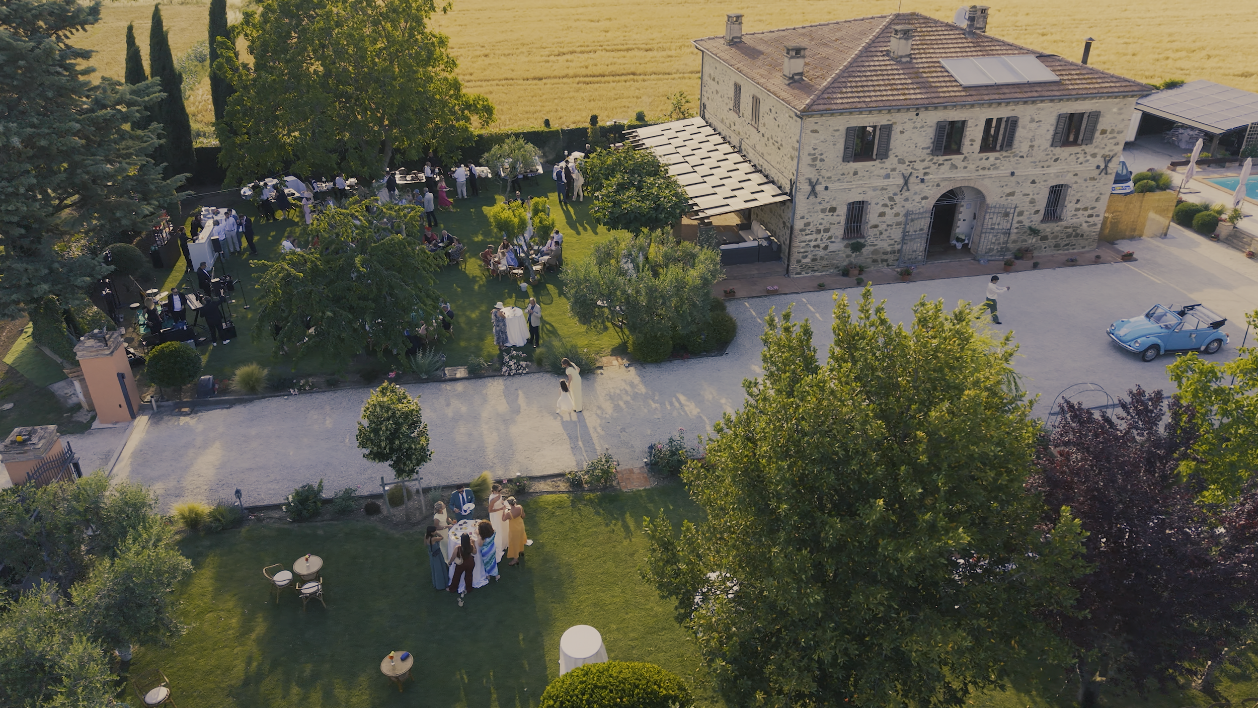 Aerial view of a celebration at a stone house with a garden, trees, and outdoor seating, with people gathered and socializing.