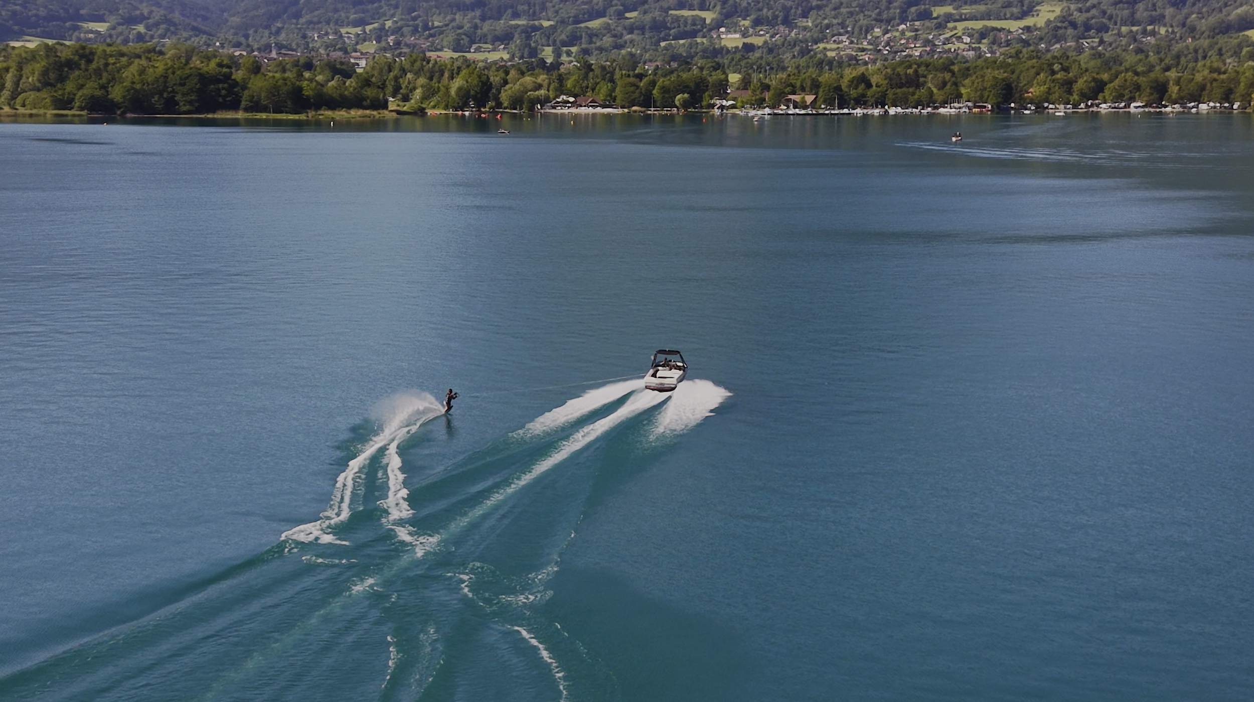 A person wakeboarding on a large, calm lake with a motorboat towing them, surrounded by green hills and trees, and a distant shoreline with houses.