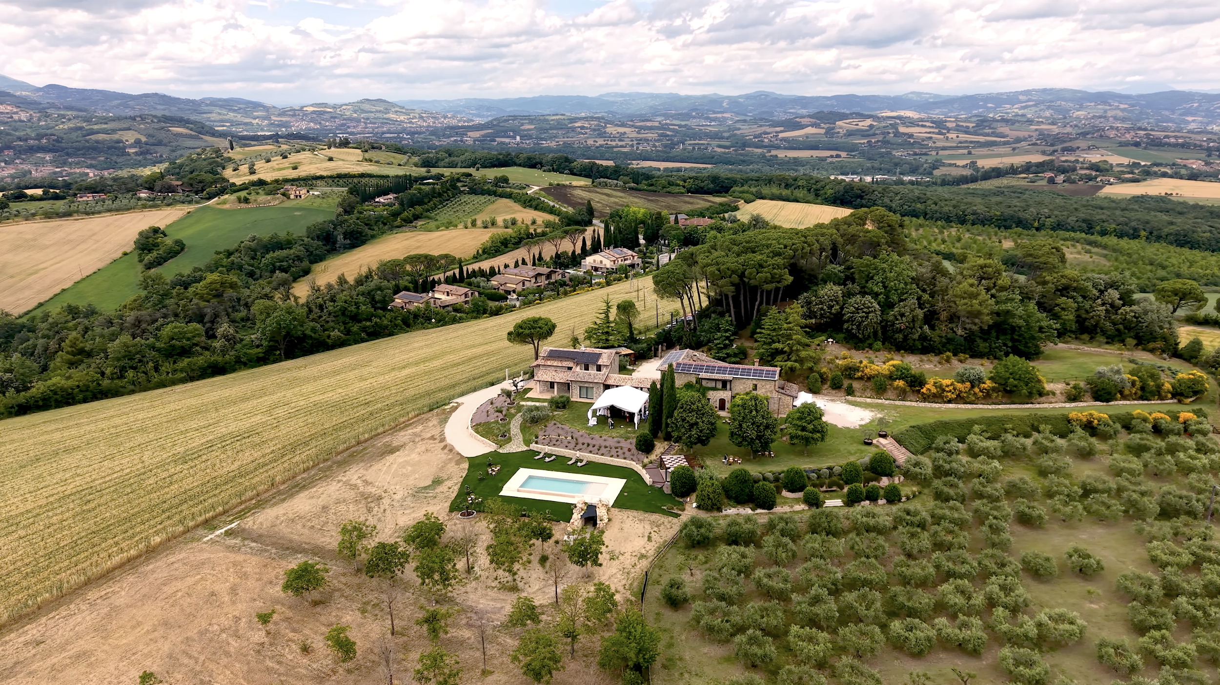 A scenic aerial view of a countryside with rolling hills, trees, and a few houses. There is a house with a swimming pool and a large garden in the foreground.