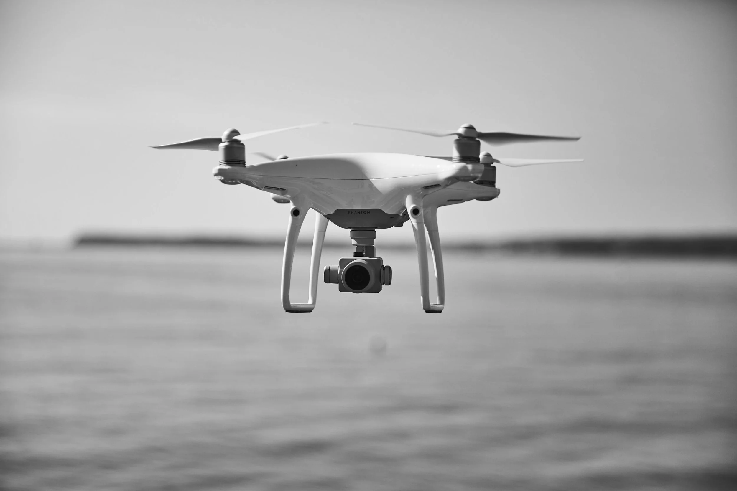 A white drone with four propellers flying outdoors, with a blurred horizon in the background, captured in black and white.