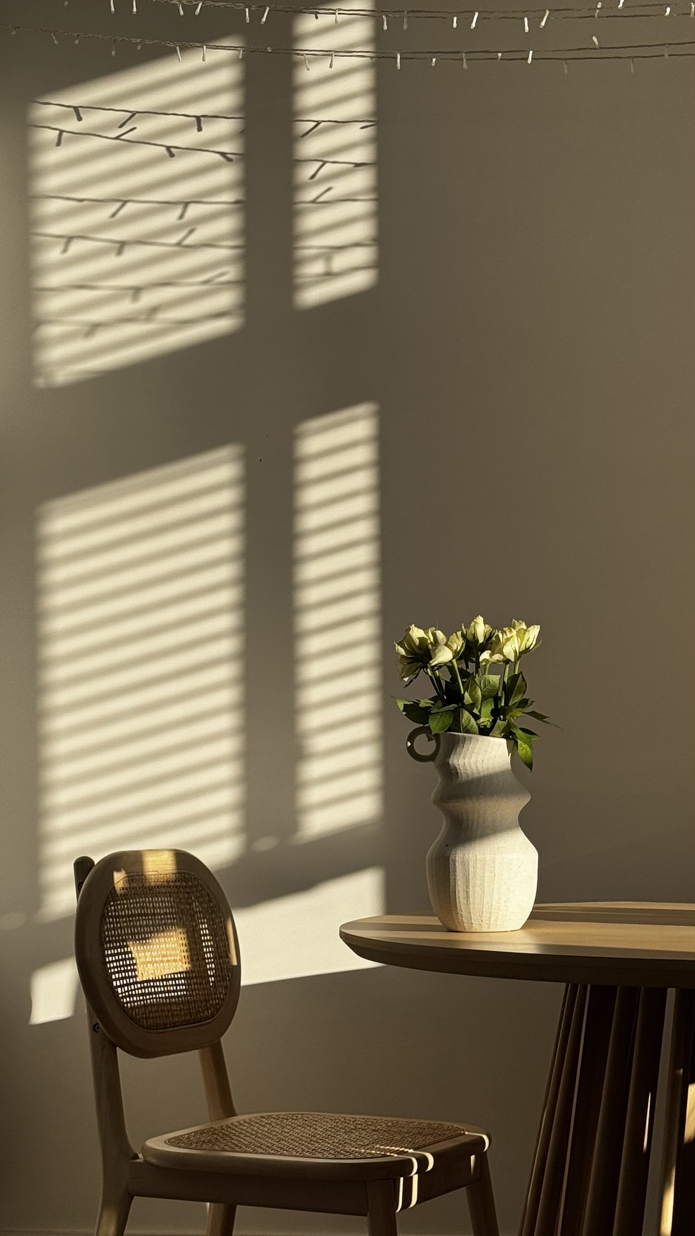 A white vase with green and white flowers on a wooden table with sunlight creating shadow patterns on the wall.