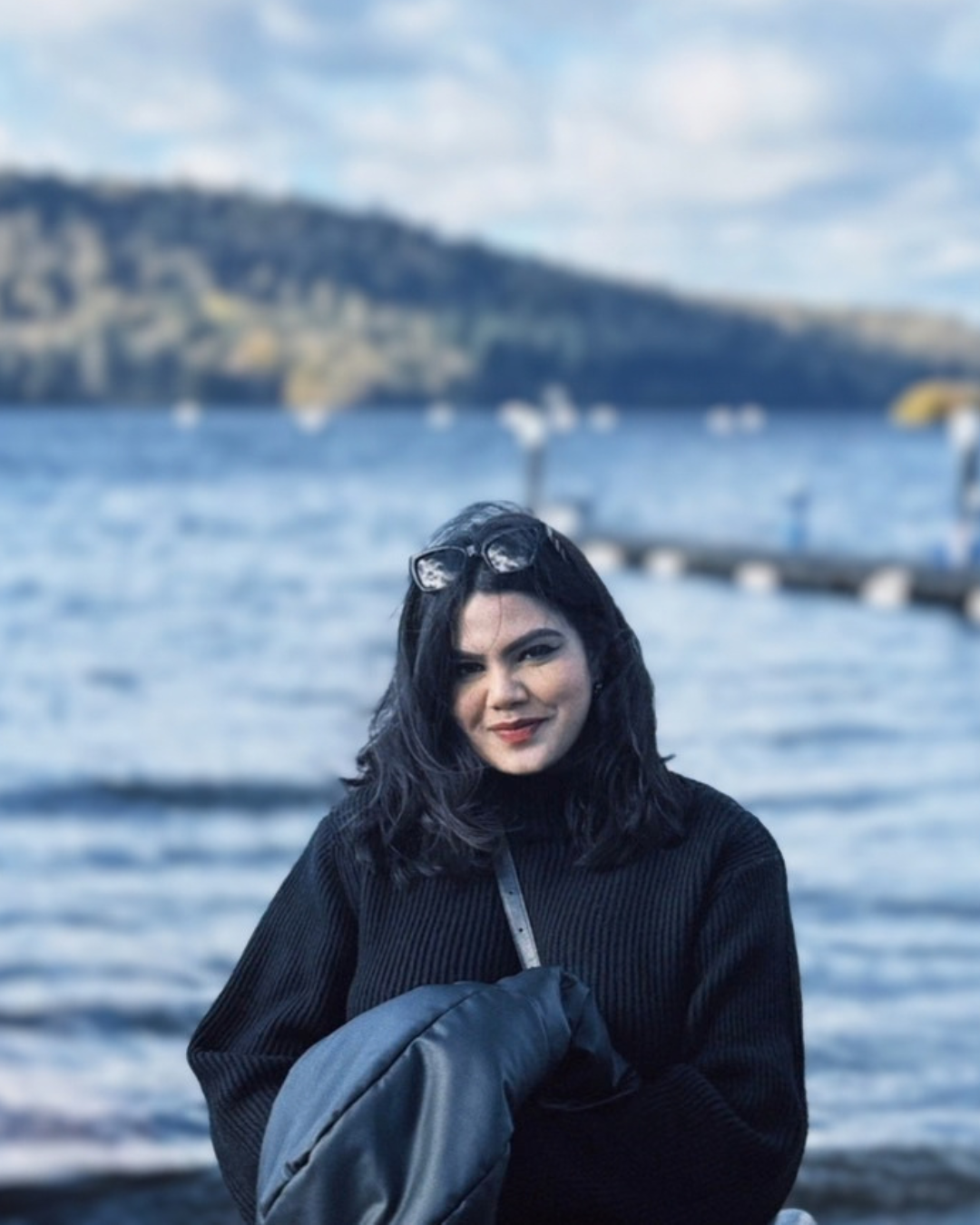 A woman with dark hair wearing a black sweater, holding a dark jacket, standing in front of a body of water with boats and hills in the background.