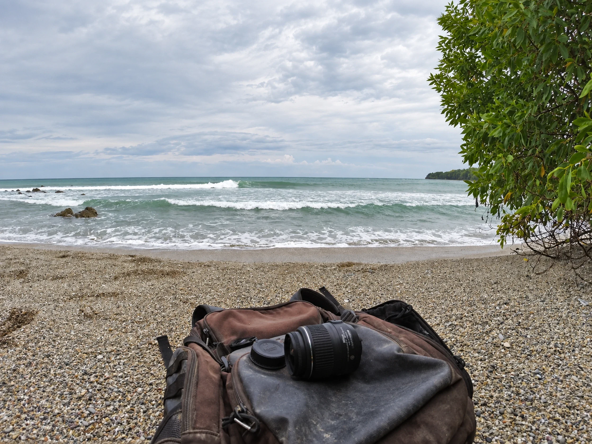 Waves breaking at Playa Cedros near Cabuya, Costa Rica, with a camera bag on the pebbled beach and trees overhead.