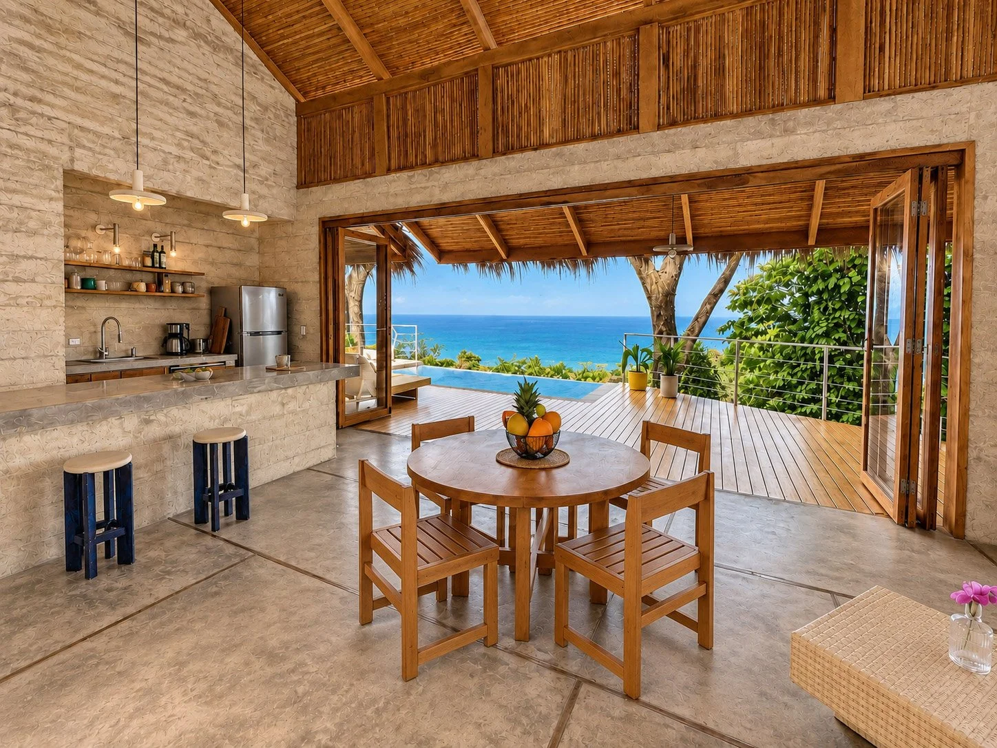 Open-concept kitchen and dining area with ocean view, featuring a round wooden table with four chairs, a fruit centerpiece, a kitchen counter with stools, open sliding doors leading to a deck, and lush greenery with a view of the ocean.