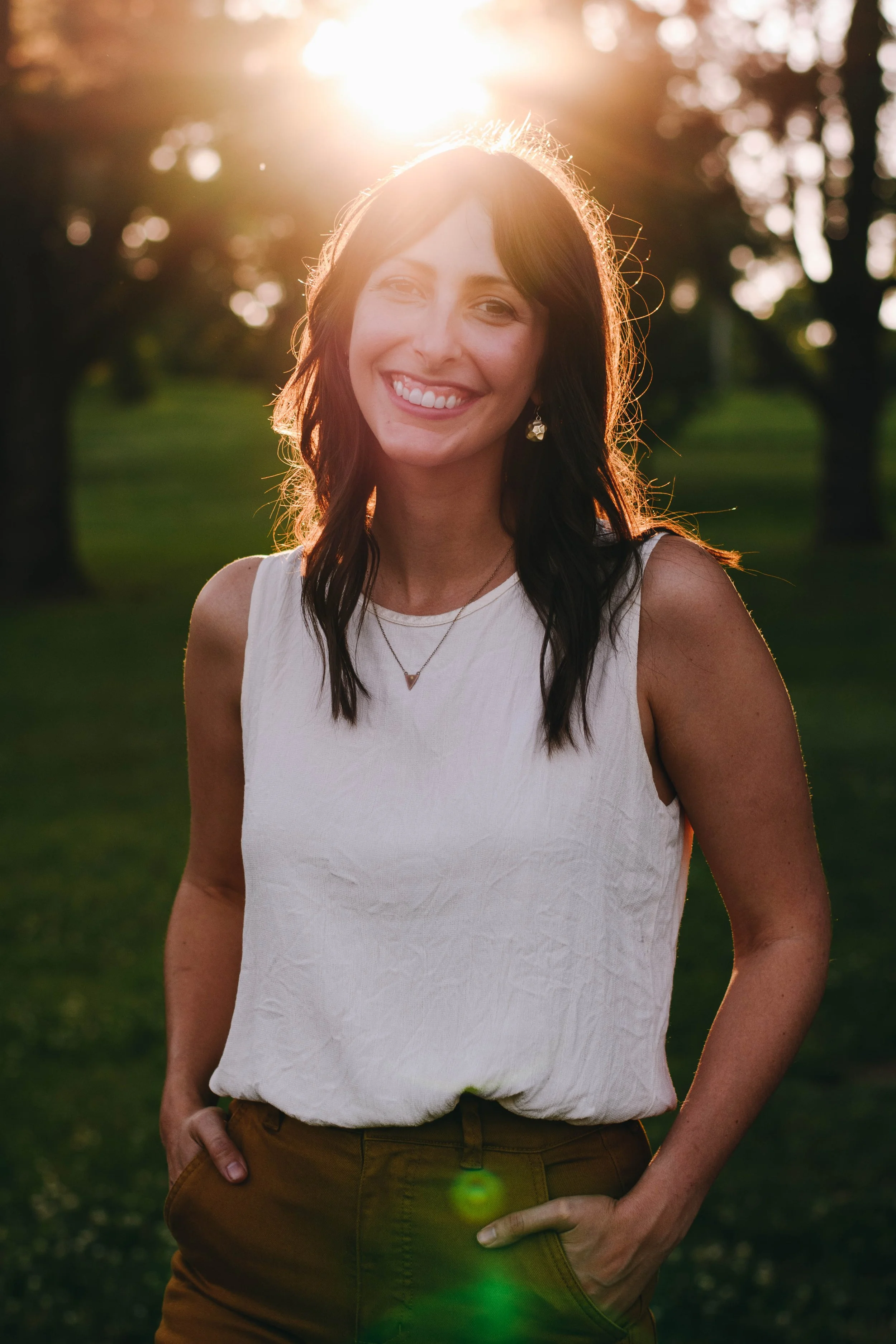 A woman smiling outdoors at sunset, wearing a white sleeveless top and jewelry, with her right hand in her pocket and natural sunlight behind her.