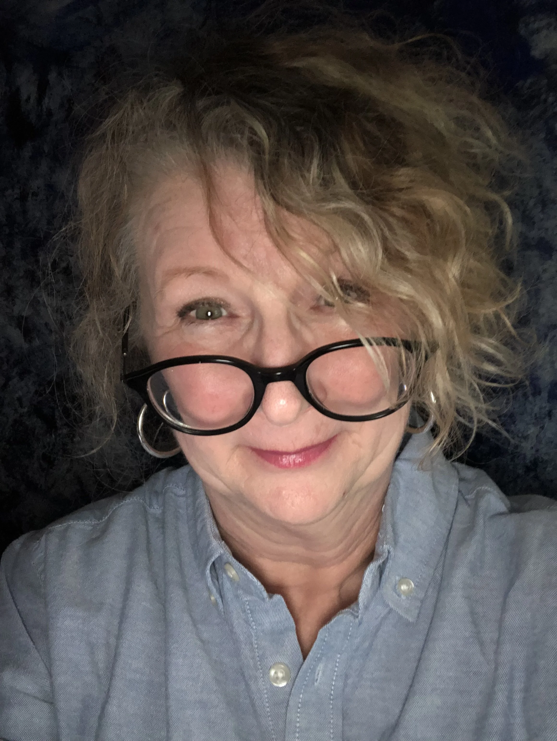Close-up of a woman with curly blonde hair and glasses, smiling and wearing a light blue shirt, lying on a dark surface.