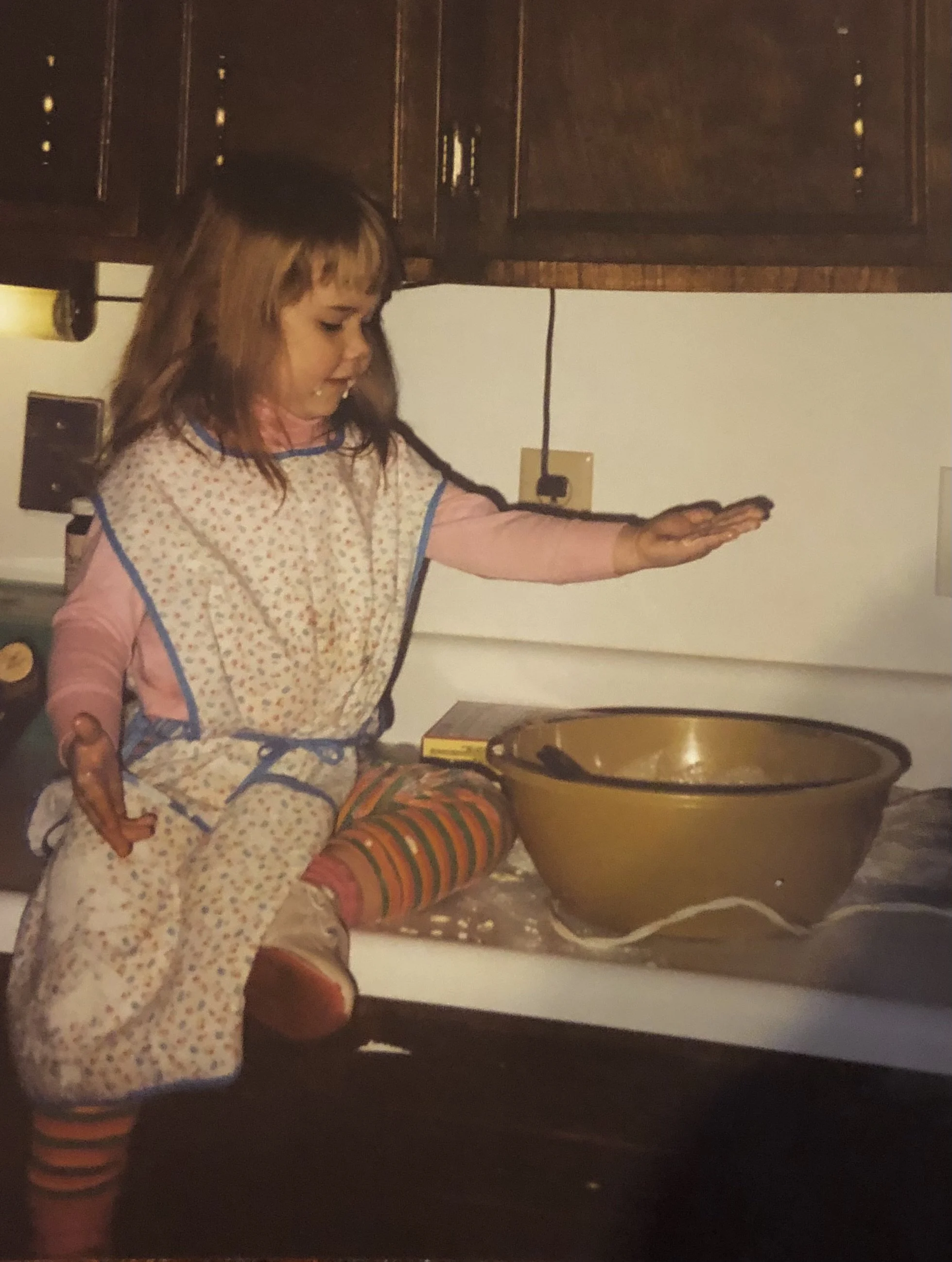 A young girl sits on a kitchen counter wearing her grandmother's apron and stirring cookie dough with a wooden spoon, capturing the joy of learning family recipes across generations.